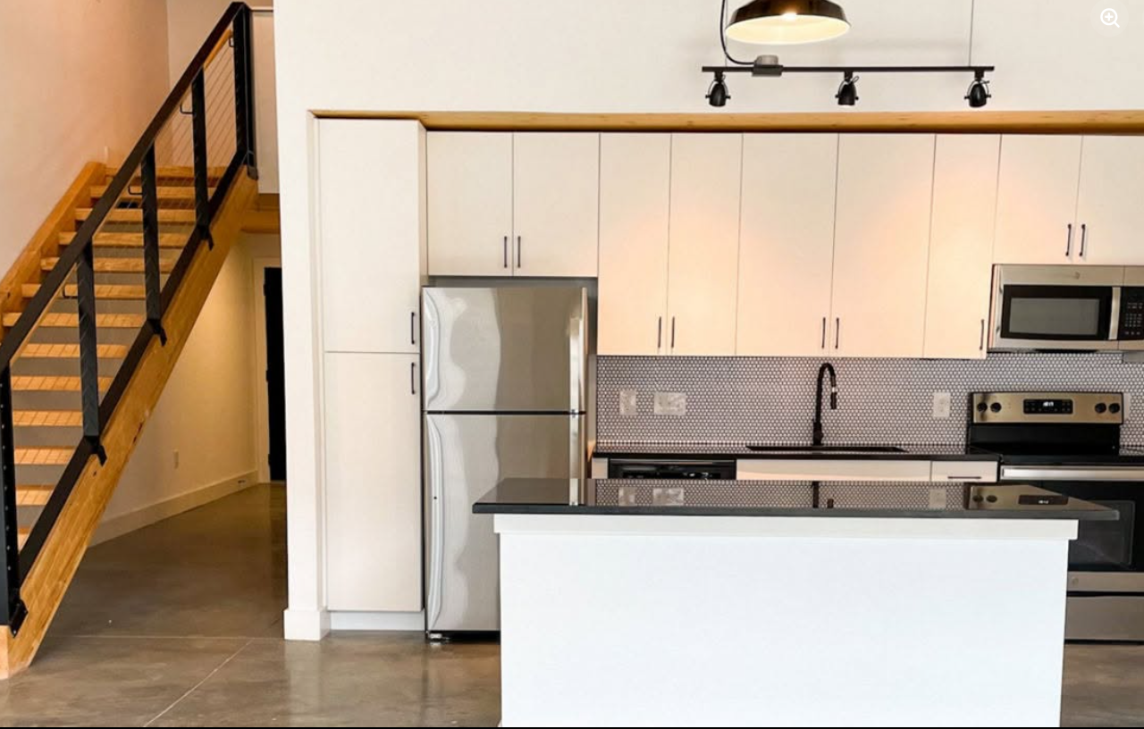 A kitchen inside a Judson Mill Lofts apartment features white cabinetry, stainless-steel appliances, a patterned backsplash and a large island, with a wooden staircase adding to the loft's industrial-style design.