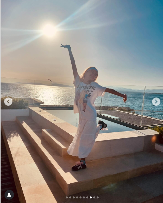 Kate Hudson was photographed with a playful pose against the golden sunset on a seaside terrace, wearing a breezy white skirt and graphic tee. The stunning coastal backdrop captured the dreamy essence of her stay in Greece | Source: Instagram/katehudson