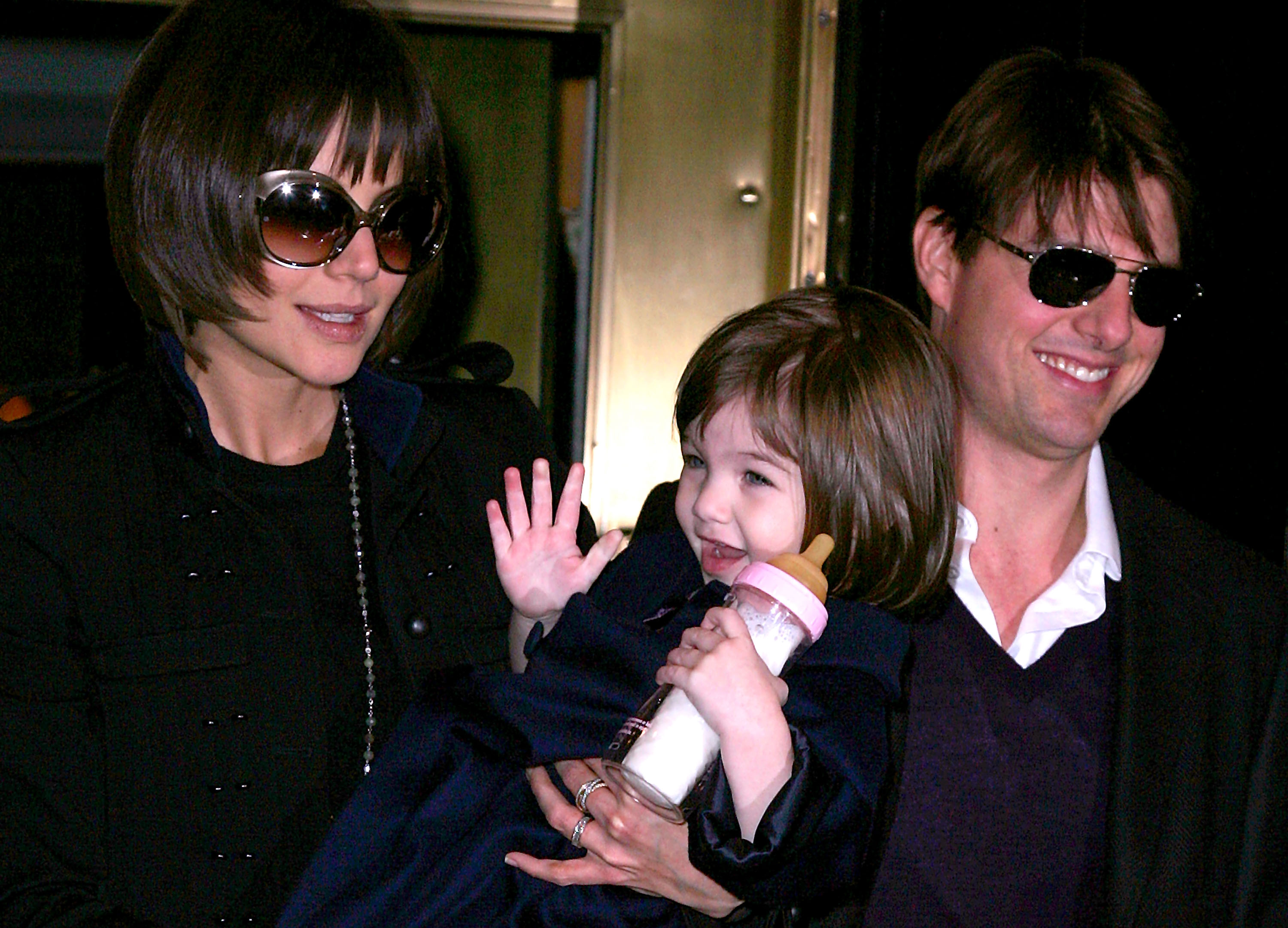 In this candid moment, baby Suri Cruise rests comfortably in Katie Holmes’ arms, clutching a milk bottle with tiny hands. Her parents, dressed in polished black ensembles, are seen stepping out for an appearance, embodying Hollywood’s most-watched family at the time. The scene is both poised and intimate—a rare glimpse into their lives behind the spotlight, with Suri quietly stealing the moment.
