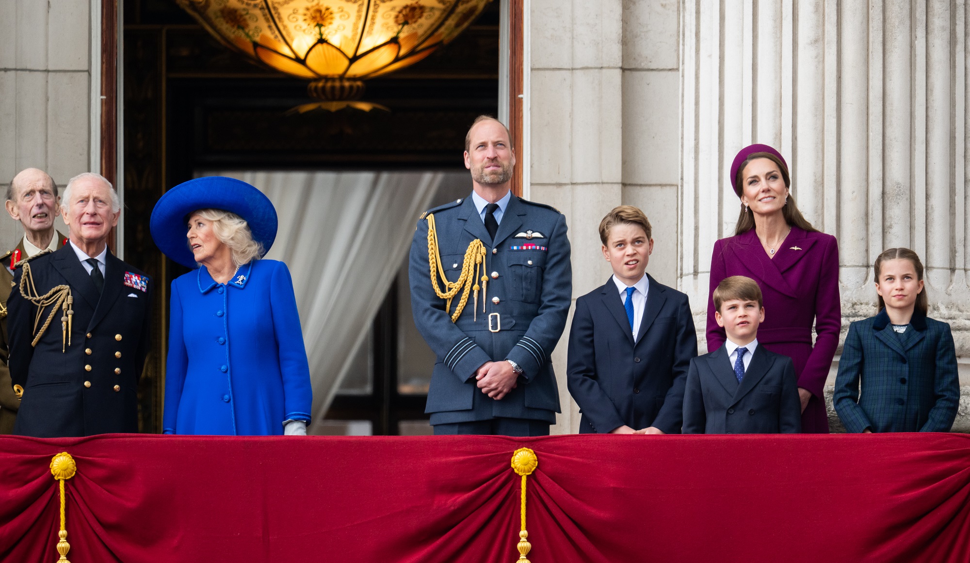 King Charles and Queen Camilla stand at the far left of the balcony, both looking skyward—he in a decorated navy uniform, she in a bold blue coat and wide-brimmed hat. Next to them are William in RAF uniform, George, Louis, and Charlotte in dark formalwear, and Catherine in deep plum.