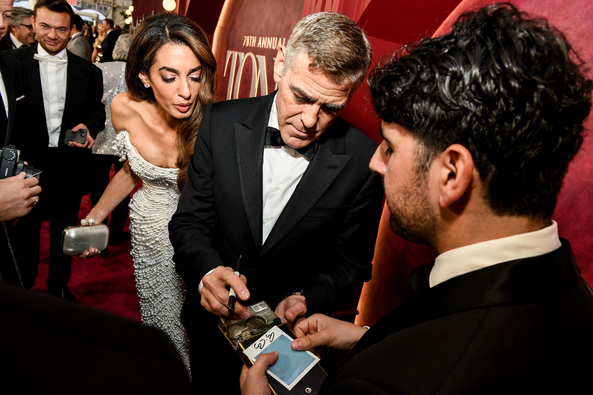 George Clooney signs memorabilia while Amal Clooney leans in to observe, her expression focused. The couple pauses amid a swirl of fans and flashing cameras on the red carpet.