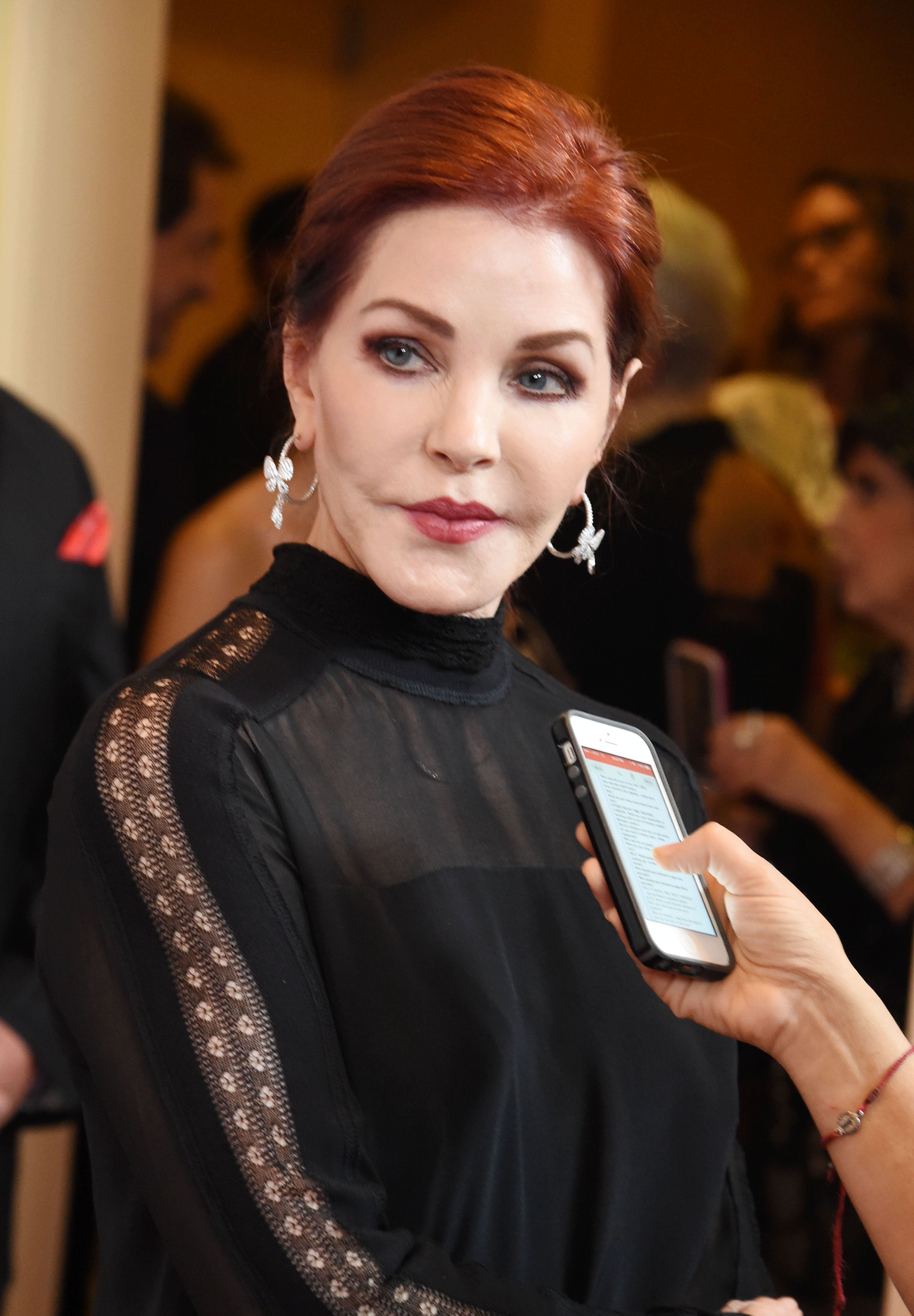Priscilla Presley pauses for an interview while attending the Last Chance for Animals Benefit Gala at The Beverly Hilton Hotel in Beverly Hills, California, on October 24, 2015, wearing a high-neck black top featuring sheer lace trim along the sleeves and chest, complemented by diamond statement earrings and her deep auburn hair styled back into a neat updo.