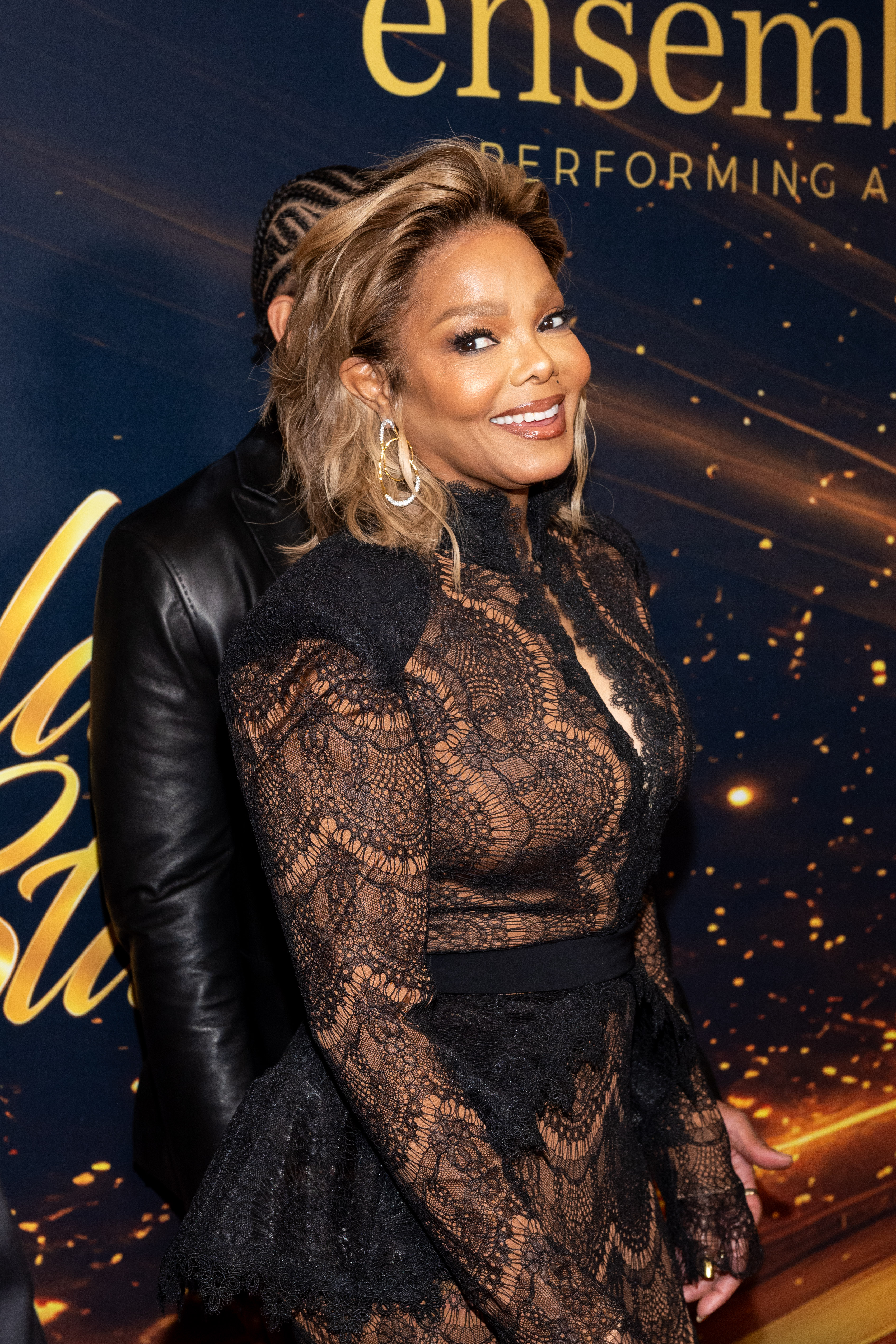 Janet Jackson smiles at the camera in her sheer black lace gown, showing off its intricate patterns, structured shoulders, and fitted waist, with a person partially visible behind her. | Source: Getty Images