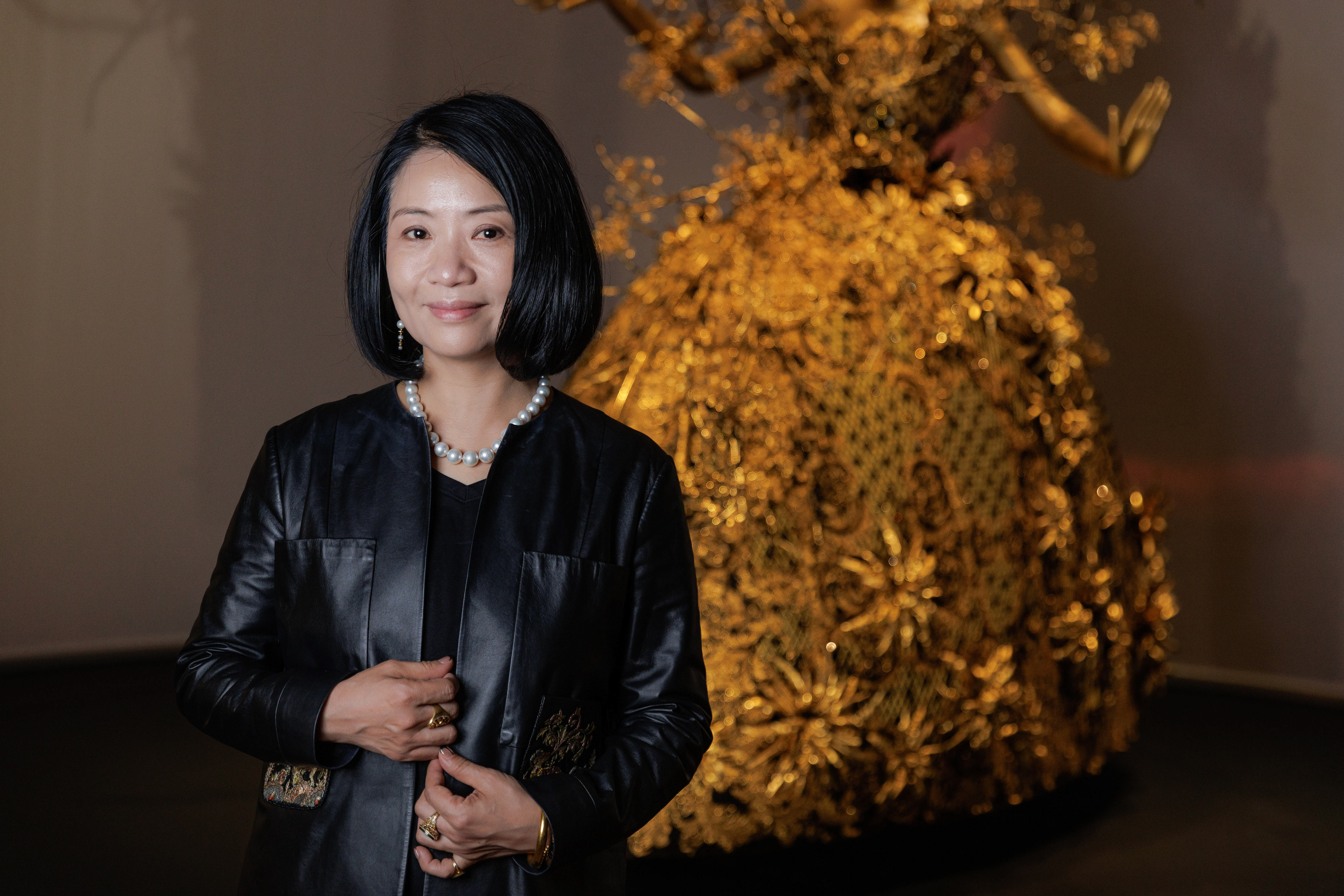 Famed haute couture designer Guo Pei posed before one of her breathtaking gold sculptural creations during her solo exhibition at the M+ Gallery on September 21, 2024, in Hong Kong, China. Embracing a sleek and modern look, she styled a pearl collar necklace over an understated black ensemble paired with an edgy black leather coat, allowing the dazzling craftsmanship behind her to take center stage. | Source: Getty Images