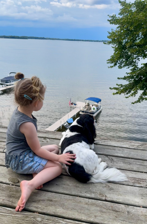 Kaliyah sits on a wooden deck with one hand resting on a black-and-white dog as they look toward the water. A dock and boats are visible below, showing another outdoor setting featured in the family photographs. | Source: Instagram/wynonnajudd