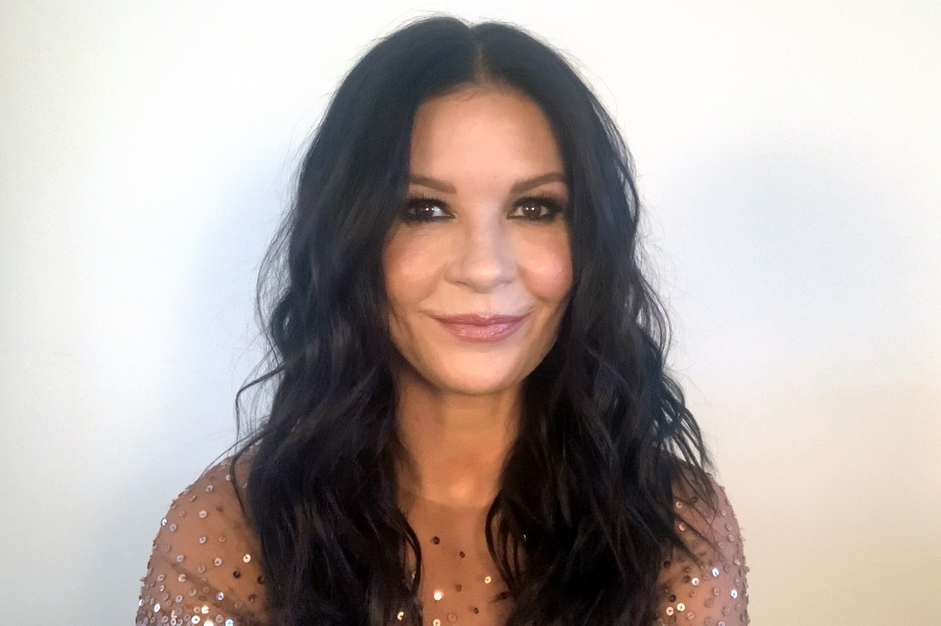 Catherine Zeta-Jones smiles warmly into the camera while appearing remotely to present during the virtual broadcast of the "26th Annual Critics Choice Awards" on March 7, 2021. She frames her soft-glam makeup and glossy pink lip with center-parted textured brunette waves, wearing a sheer, skin-toned evening top embellished with scattered shimmering copper sequins.