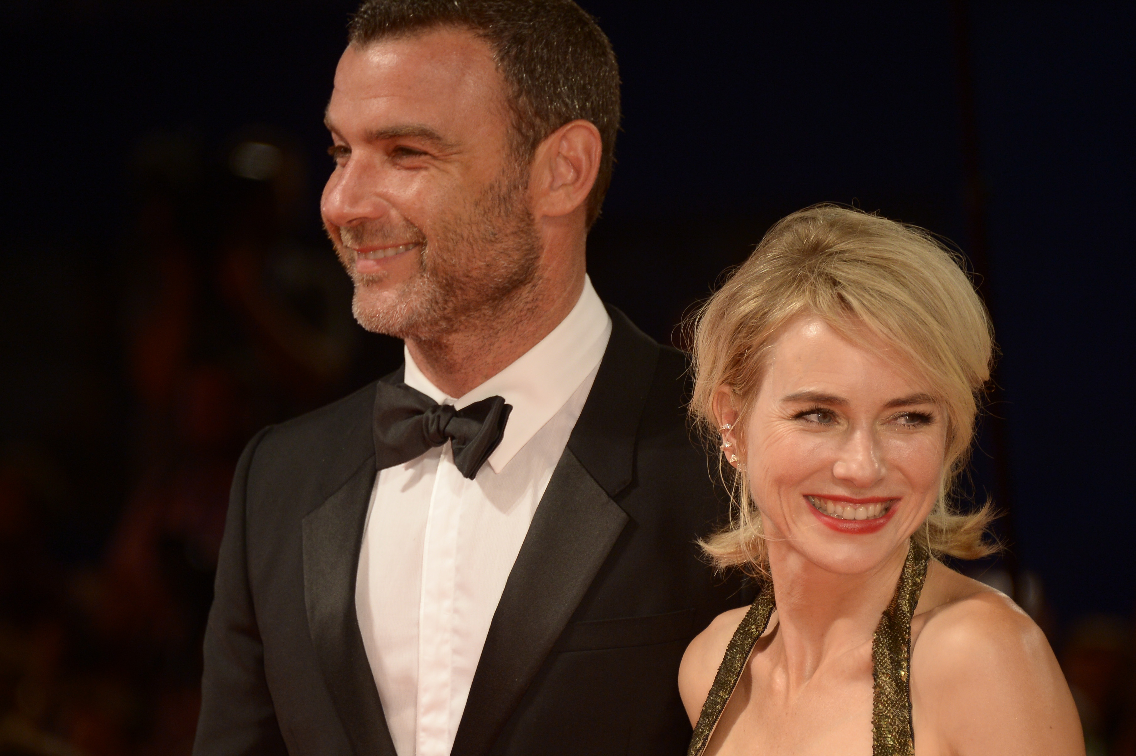 Naomi Watts and Liev Schreiber walk the red carpet at a glamorous event. Watts dazzles in a shimmering halter gown while Schreiber wears a classic black tuxedo. Both smile warmly as they pose together under the bright event lights.
