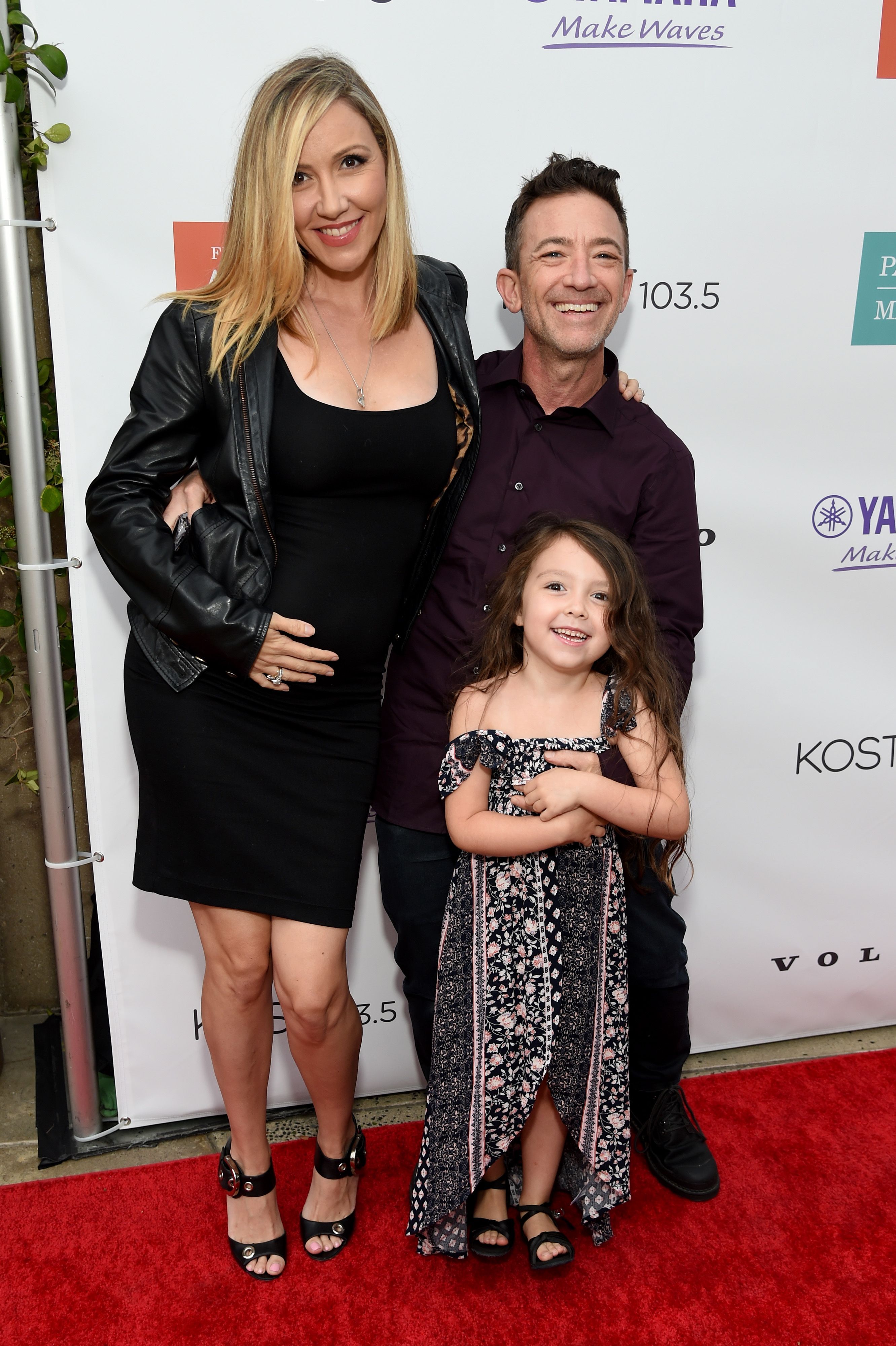 David Faustino smiled alongside his partner, Lindsay Bronson, and their daughter, Ava Marie Grace Faustino, while attending the Festival of Arts Celebrity Benefit Event on August 28, 2021, in Laguna Beach, California. The family posed together on the red carpet, with Bronson cradling her baby bump in a fitted black dress and leather moto jacket, Faustino sporting a deep purple button-down shirt, and young Ava beaming in a floral off-the-shoulder maxi dress. | Source: Getty Images