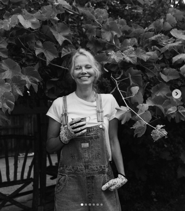 In a luminous black-and-white portrait, Pamela Anderson flashes a radiant, makeup-free smile against a dense wall of grapevine leaves at Arcady. Wearing denim overalls, garden gloves, and holding a small teacup, she exudes pure joy while reflecting on "Pamela's Cooking with Love," calling the Vancouver Island show a true dream project that blends her flourishing garden with international culinary talent. | Source: Instagram/pamelaanderson