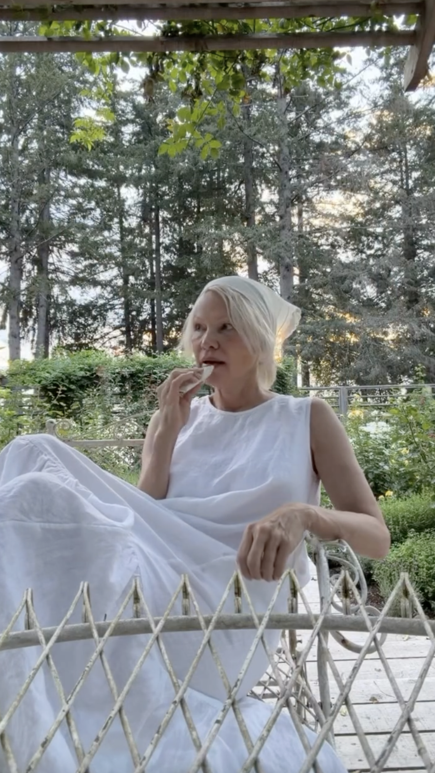 Pamela Anderson lounges peacefully in a vintage garden chair at Arcady, wrapped in a tiered white sleeveless dress with a cream headscarf tied around her blonde hair. Gazing into the distance while casually enjoying a bite to eat, she embodies the tranquil, grounding pace of estate life tucked away among the tall pines of British Columbia, sharing the moment with the caption, "Live romantically wherever you are… 🤍🌷". | Source: Instagram/pamelaanderson