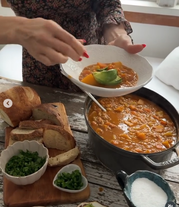 Pamela Anderson prepares a hearty homemade vegetable stew inside her cozy Vancouver Island kitchen, garnishing a white bowl with fresh avocado slices directly from a rustic wooden dining table. Surrounded by crusty sliced sourdough bread, chopped cilantro, diced peppers, and flaky sea salt, the plant-based spread reflects her passion for nourishing, garden-to-table meals. She paired the cooking video with the inspiring caption, "See the beauty in who you are, then share it. 🤍". | Source: Instagram/pamelaanderson