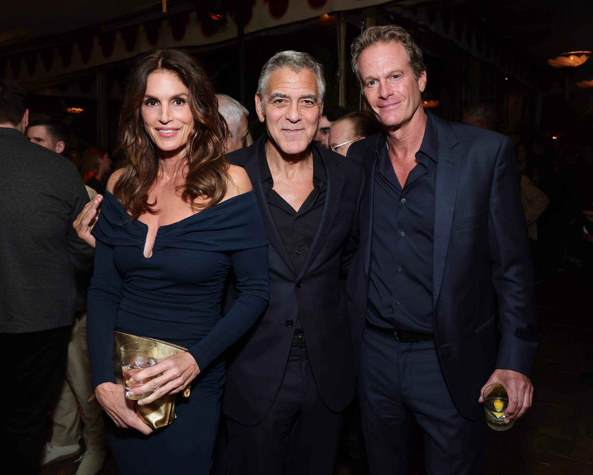 Longtime friends Cindy Crawford, George Clooney, and Rande Gerber came together for the Los Angeles premiere of "Jay Kelly" at Chateau Marmont on November 11, 2025. Posing closely with drinks in hand, Cindy stunned in an off-the-shoulder navy ruched long-sleeve dress paired with a metallic clutch, flanked by Clooney and husband Rande, who both looked dapper in dark tailored suits. | Source: Getty Images