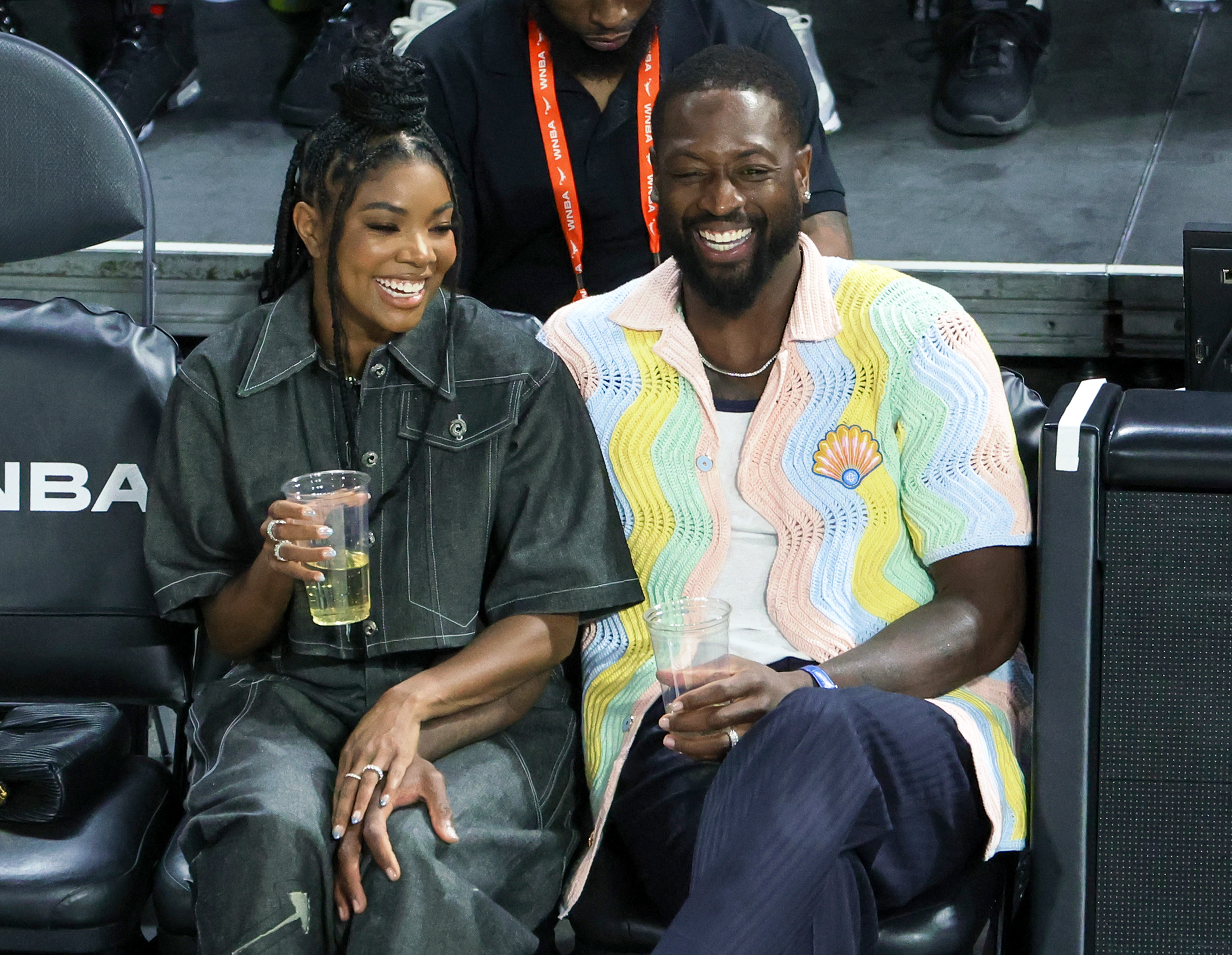 The couple appears again courtside, this time sharing a candid, joyful moment. Gabrielle, in a dark outfit with her hair styled up, laughs while holding a drink. | Source: Getty Images