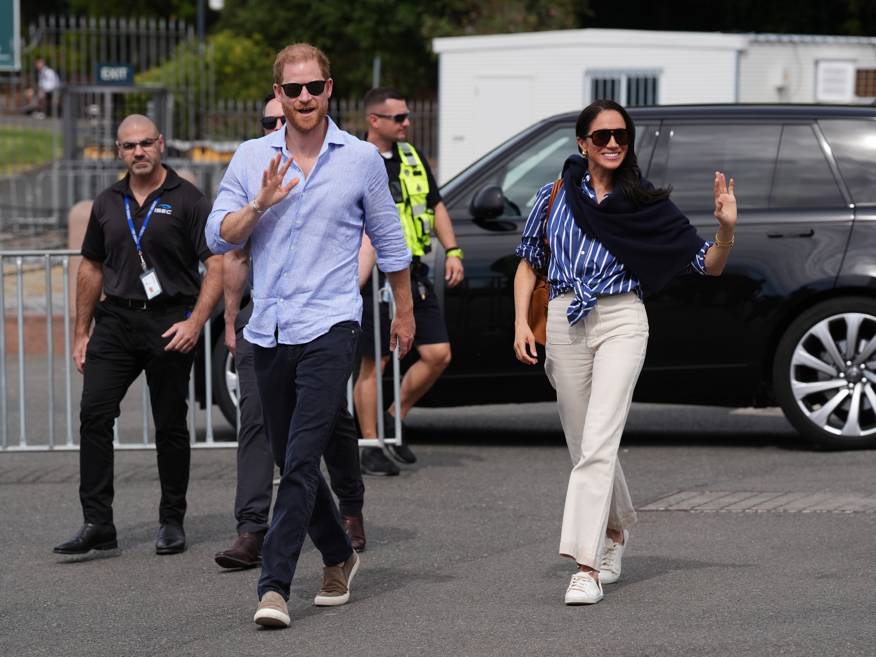 Meghan, Duchess of Sussex, waves to well-wishers alongside Prince Harry as they arrive at the Man O'War Steps in Sydney, Australia, on April 17, 2026, ahead of a sailing event with Invictus Australia members, wearing a blue-and-white striped button-down tied at the waist with off-white wide-leg trousers, sunglasses, a dark knit draped over her shoulders, and white low-top sneakers. | Source: Getty Images
