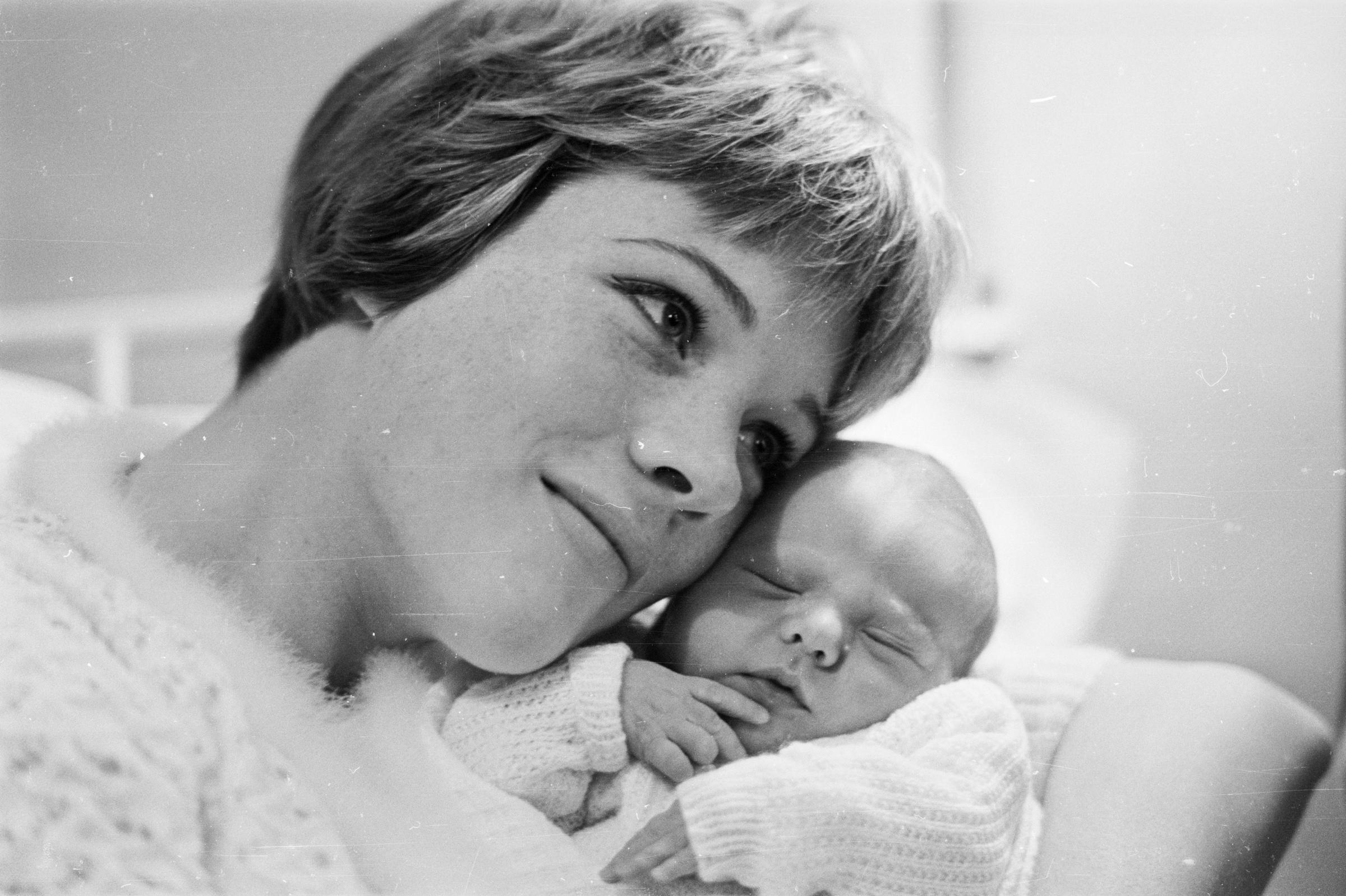 Julie Andrews cradles her sleeping newborn daughter, Emma Walton Hamilton, resting her cheek lovingly beside the infant in an intimate black-and-white portrait taken on November 28, 1962. The tender snapshot highlights the new mother's soft pixie haircut and warm expression as her baby rests peacefully swaddled in a knitted white sweater.