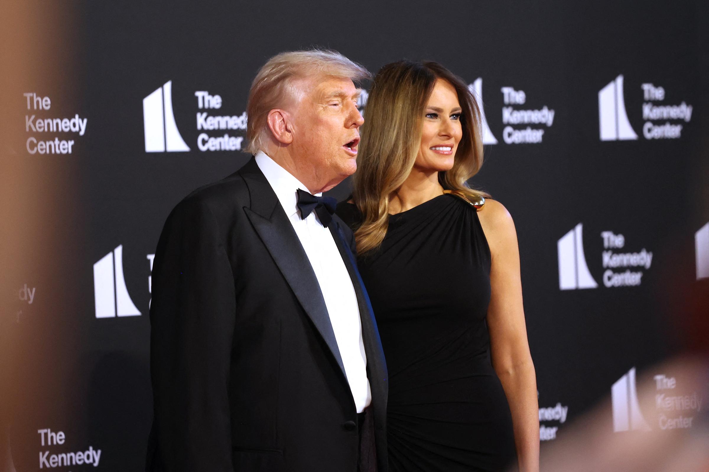 Framed by a crowd of onlookers, the president speaks as the first lady smiles beside him, both facing cameras during their arrival at the Kennedy Center's red carpet event.