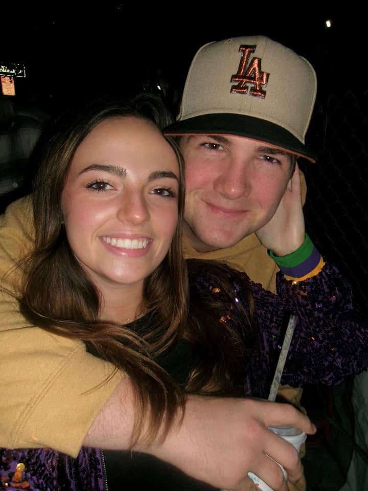 Leah Victoria Fonte and Shane David Hirstius share a warm embrace during an evening outing in a sweet, candid snapshot. Leah smiles brightly toward the camera wearing a black top and a sparkly purple sequined jacket, while Shane wraps both arms snugly around her from behind, sporting a tan hooded sweatshirt, a two-tone baseball cap, and a festive wristband while holding a drink. | Source: Facebook/Leah Fonte