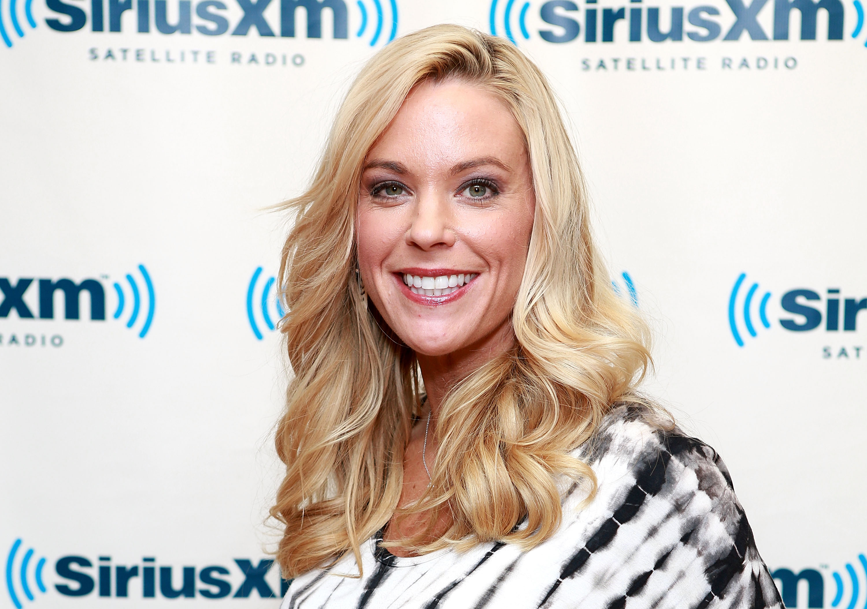 At SiriusXM Studios in New York, Kate Gosselin flashes a wide smile in front of the logo wall. Her long blonde waves and black-and-white top pair with glowing makeup, giving her a confident, camera-ready look.