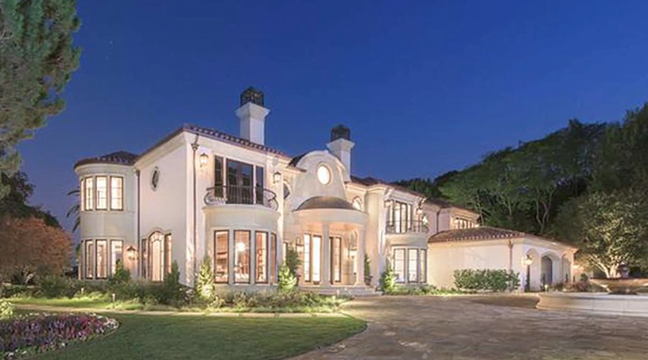 The exterior facade of Reba McEntire's former Beverly Park estate in Beverly Hills, California, glows at dusk, showcasing its Mediterranean-style architecture, off-white stucco walls, terracotta tile roofing, arched windows, and an expansive cobblestone motor court complete with a stone fountain and manicured landscaping. | Source: YouTube/Taste of Country