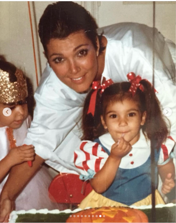 A young Kris Jenner poses with daughters Kourtney and Kim Kardashian at a family birthday celebration, with Kourtney standing in front of her beside a birthday cake. Kris wears a white top while the girls are dressed in colorful outfits. | Source: Instagram/krisjenner