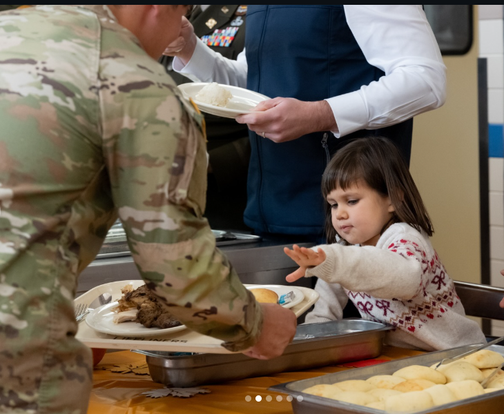 Mirabel Vance reaches toward a serving tray as a uniformed service member places food on a plate, while JD stands nearby holding a dish. | Source: Instagram/vp