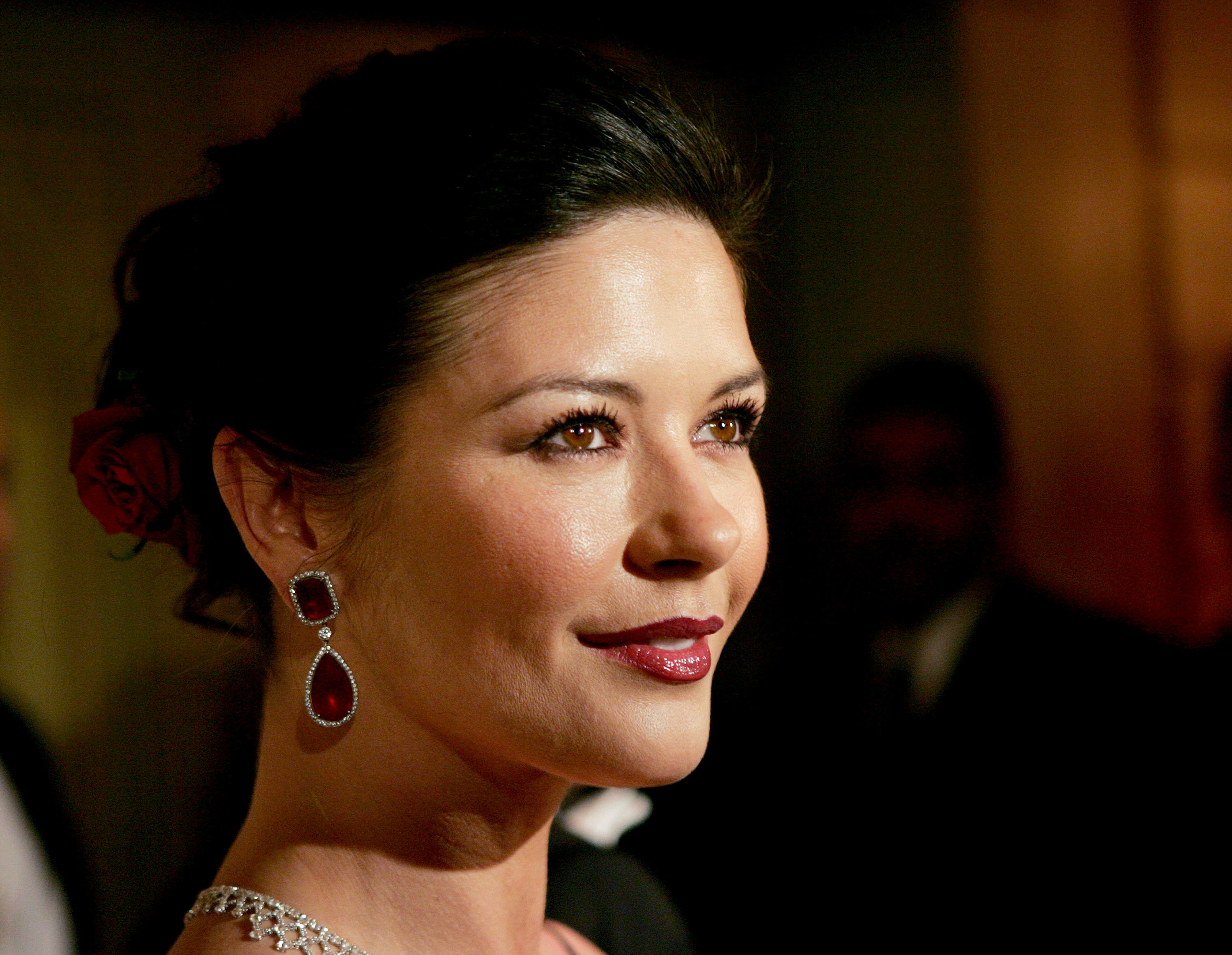 Catherine Zeta-Jones smiles warmly in profile at the Eighth Annual Red Ball at The Pierre Hotel on February 7, 2005, in New York City. She wears an elegant dark evening gown accessorized with dramatic teardrop ruby-and-diamond earrings and a coordinating diamond lattice necklace fringed with rubies, her dark hair swept up with a red rose tucked into the chignon.