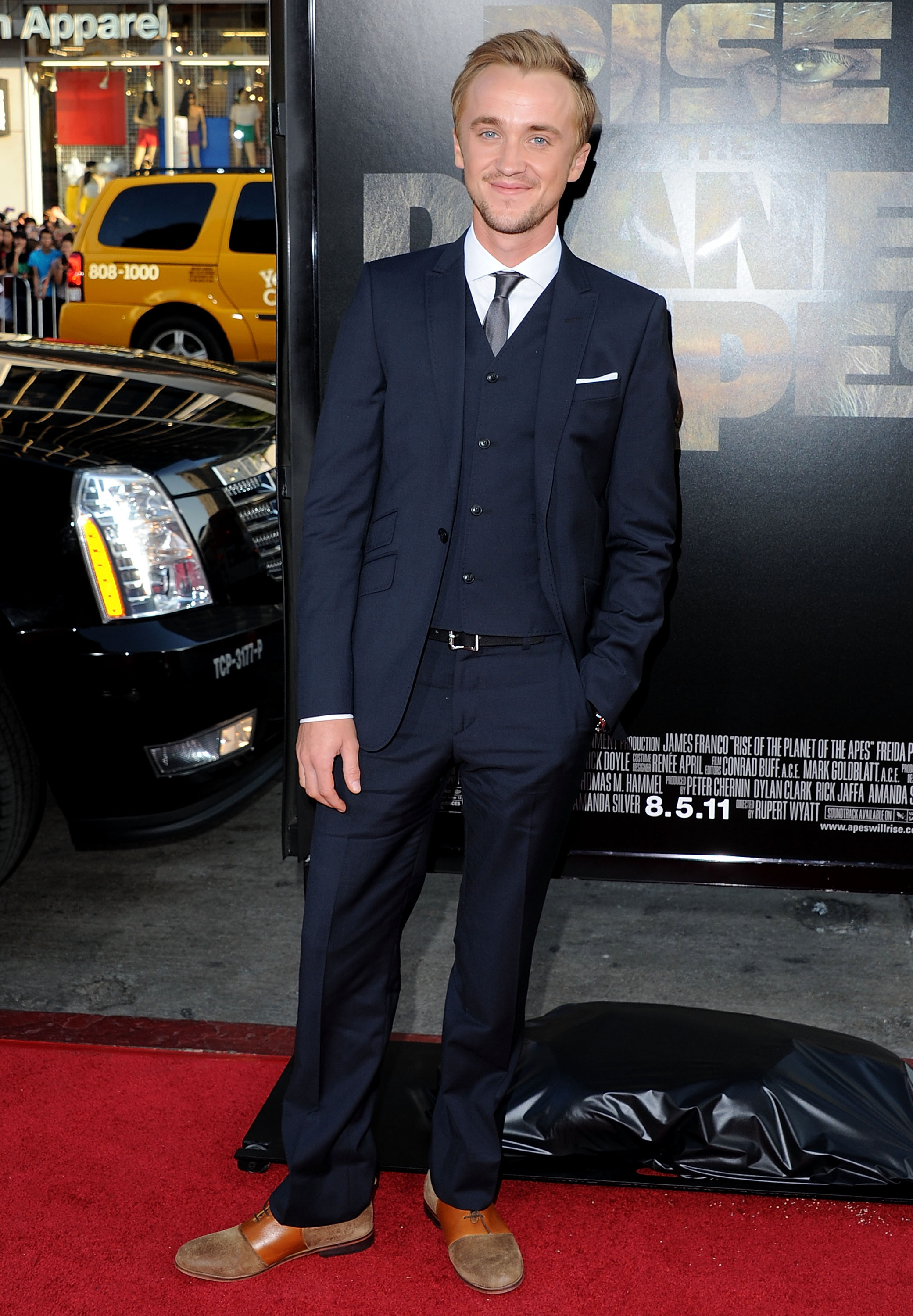 Tom Felton steps onto the red carpet in a sleek navy suit, looking effortlessly polished against the Hollywood buzz. With a calm smile and hands casually at his sides, he brings a touch of understated charm to the high-energy premiere. Set before the bold backdrop of the "Rise Of The Planet Of The Apes" poster, he stands poised at the intersection of elegance and excitement.
