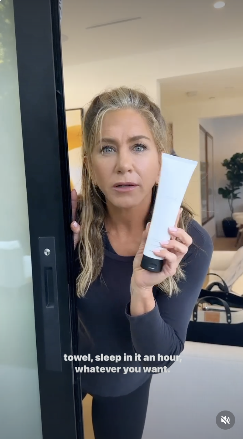 Jennifer Aniston shows off gray strands along her hairline while holding a bottle of LolaVie's Intensive Repair Treatment in a June 2023 Instagram video. The actress wears a dark long-sleeved top with her blonde hair styled half-up.