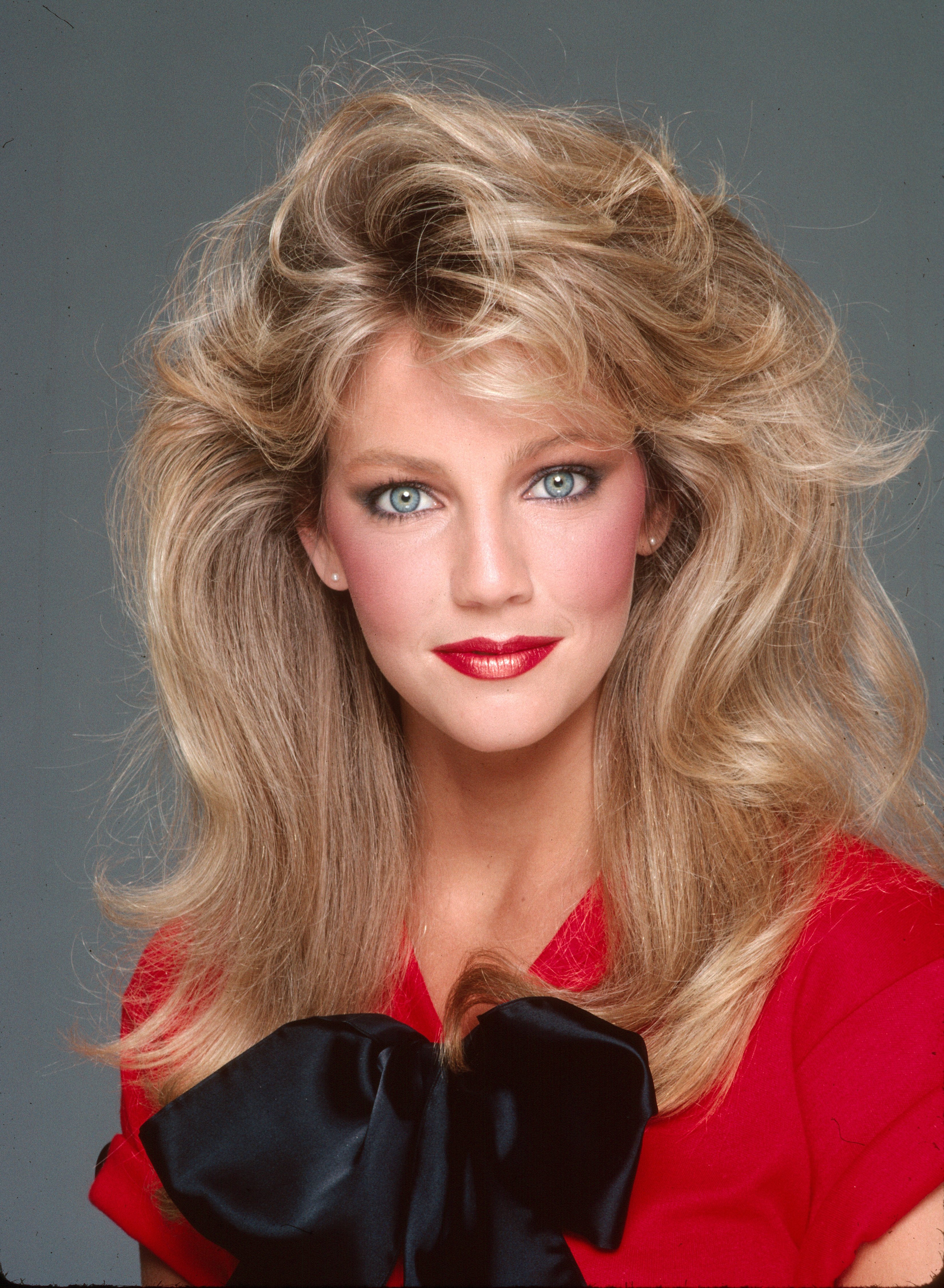 Heather Locklear epitomizes 1980s glamour in a studio portrait session from February 2, 1981, in Los Angeles, California. With voluminous, layered blonde waves framing her face, she gazes directly at the camera showcasing rosy blush, bold red lipstick, and pearl stud earrings while wearing a vibrant red top accented with an oversized black satin bow. | Source: Getty Images