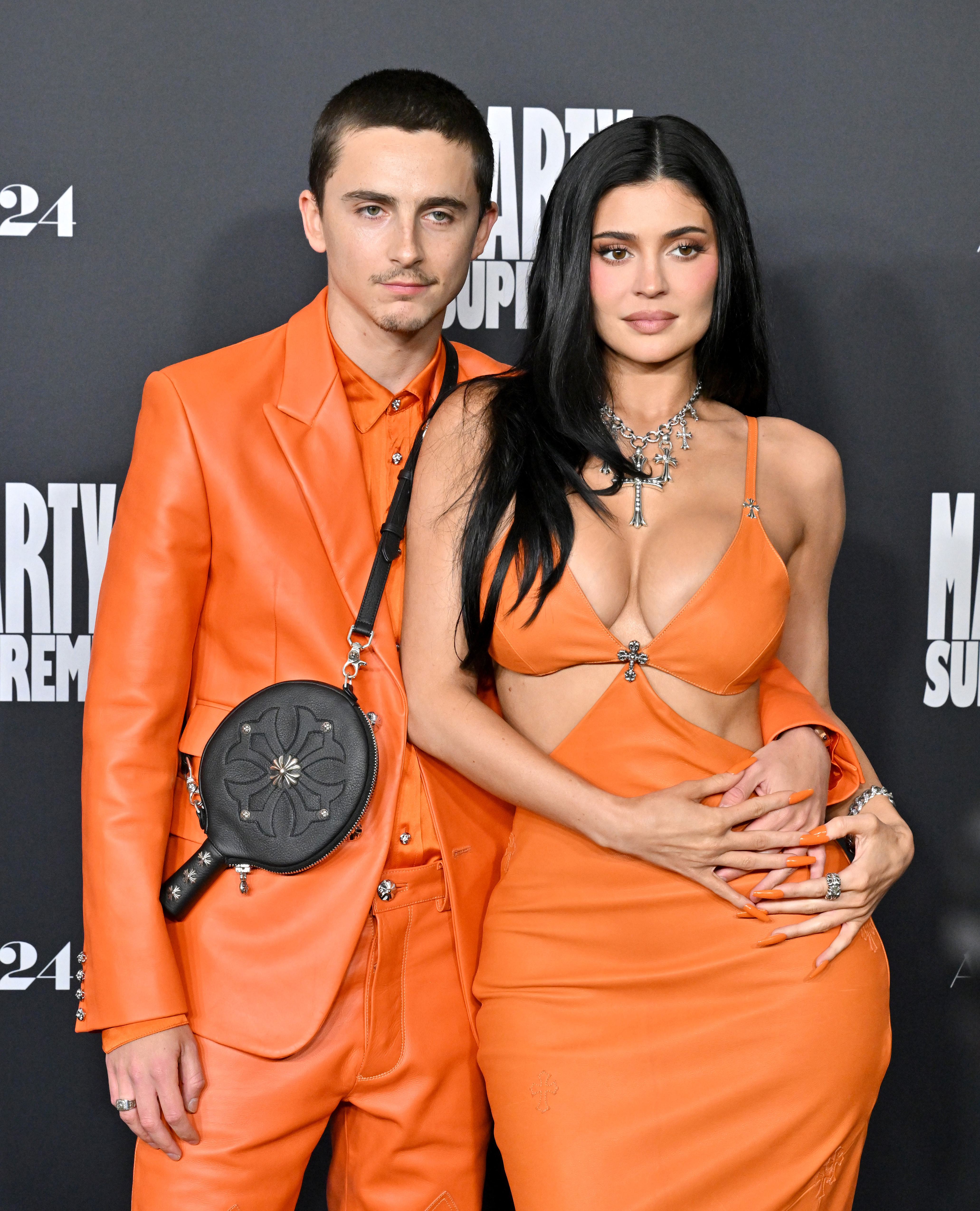 Kylie Jenner and Timothée Chalamet attend the Los Angeles premiere of Marty Supreme on December 8, 2025, wearing matching custom orange Chrome Hearts outfits. Jenner paired her cutout gown with a silver cross necklace, while Chalamet carried a black leather ping-pong paddle case referencing his role in the film. | Source: Getty Images.