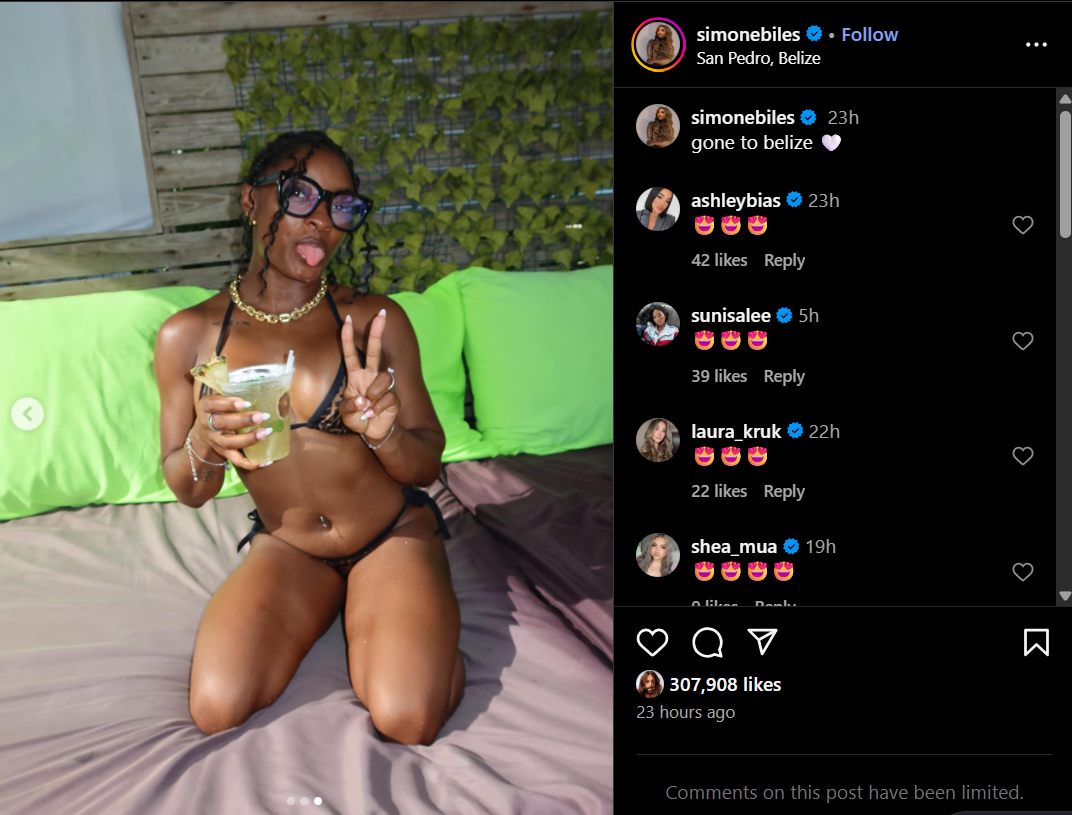 Simone Biles in a black and brown bikini, sitting in bed holding a drink and flashing a peace sign in San Pedro, Belize. From an Instagram post dated July 7, 2025. | Source: Instagram/simonebiles