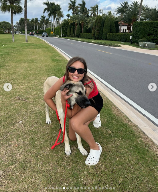 Embracing a sunny, casual Florida afternoon, a teenage Arabella knelt on a palm-lined neighborhood lawn to hug a dog. Wearing dark sunglasses, a red tank top, black shorts, and white clogs, she flashed a warm smile while holding onto the pup's bright red leash. | Source: Instagram/ivankatrump