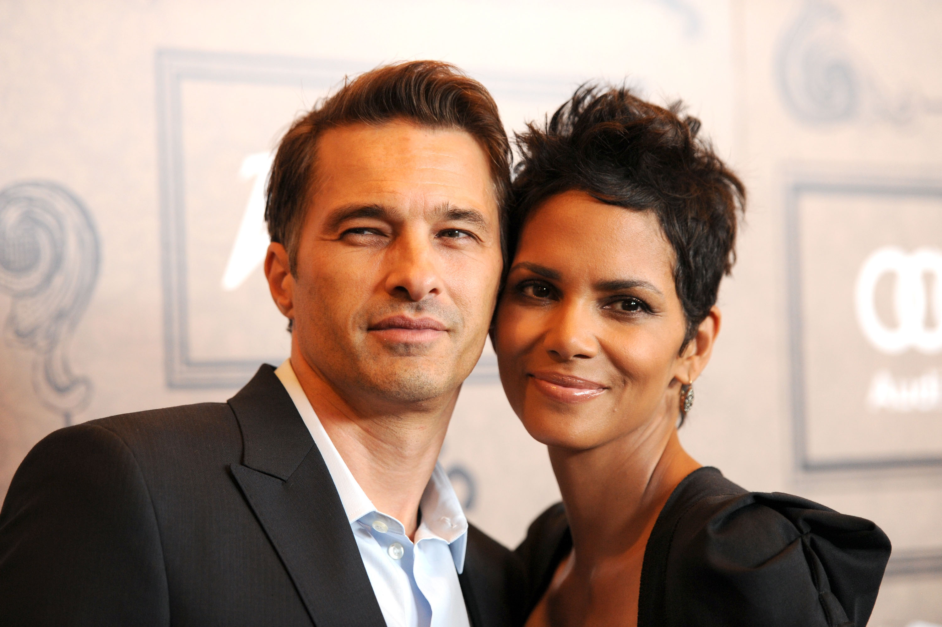 Actors Olivier Martinez and Halle Berry leaned in close as they arrived at Variety's 4th Annual Power of Women Event Presented by Lifetime at the Beverly Wilshire Four Seasons Hotel on October 5, 2012, in Beverly Hills, California. Berry showed off her textured cropped pixie cut alongside subtle drop earrings and a black puff-shoulder ensemble, smiling alongside Martinez, who wore a black tailored suit jacket over an open-collar light blue dress shirt. | Source: Getty Images