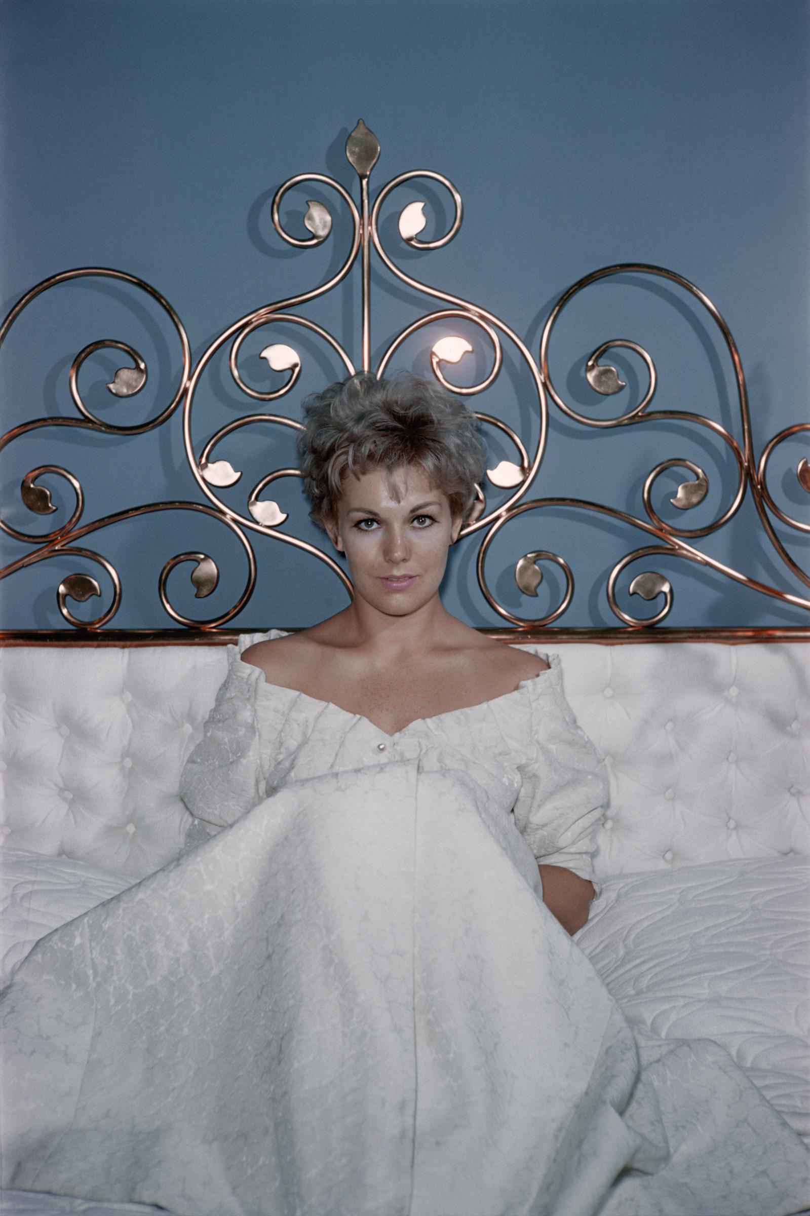 Kim Novak sits on a bed in a white off-the-shoulder dress, posing against an ornate metal headboard. | Source: Getty Images