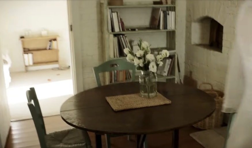 A quiet corner of the home holds a rustic round table topped with fresh flowers, nestled beside built-in shelving and a painted brick hearth — ideal for reading or morning tea. From a video dated July 20, 2015. | Source: YouTube/Vogue