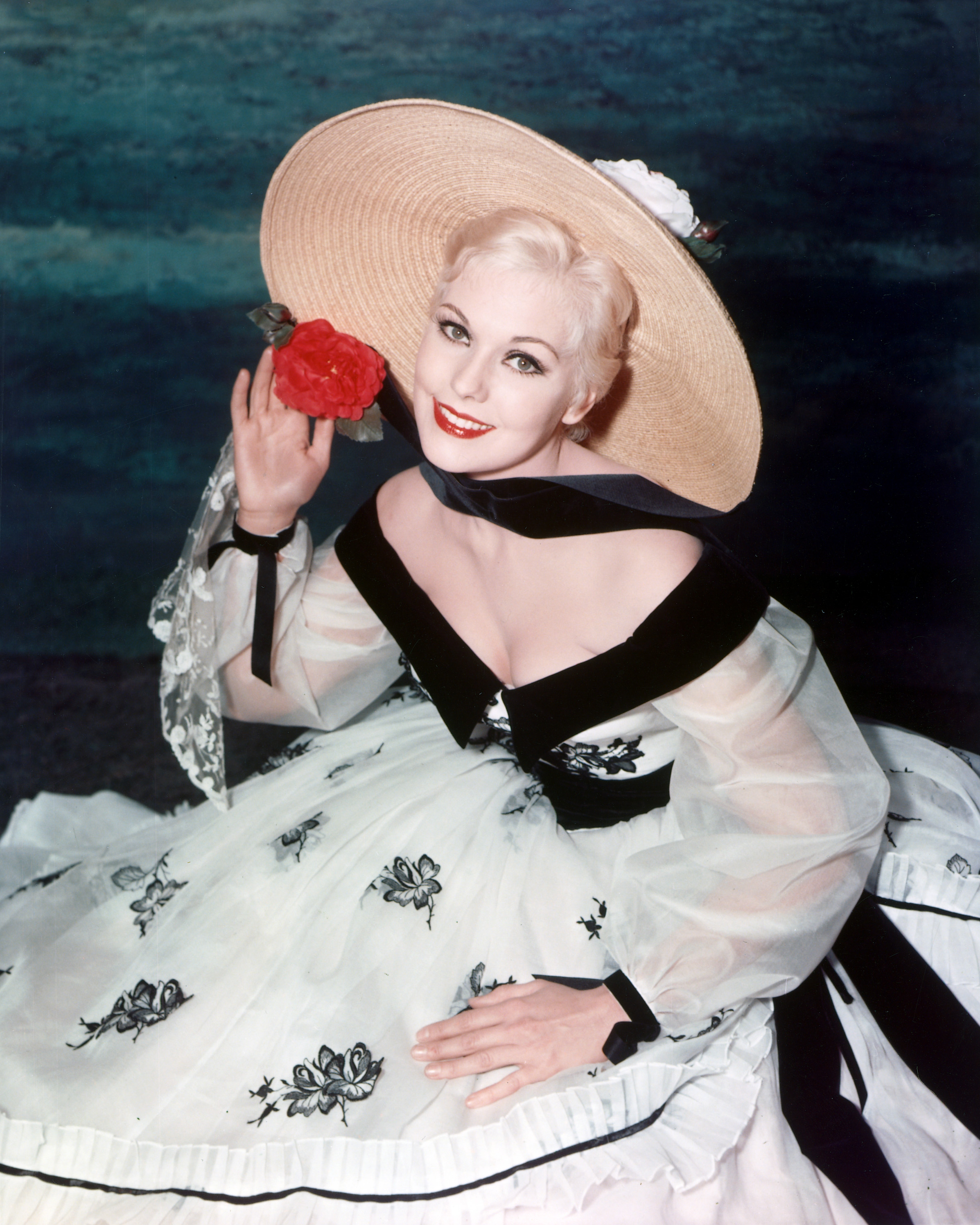 Kim Novak smiles in a studio portrait wearing a white dress with black floral details and a wide-brimmed straw hat adorned with a red flower. | Source: Getty Images