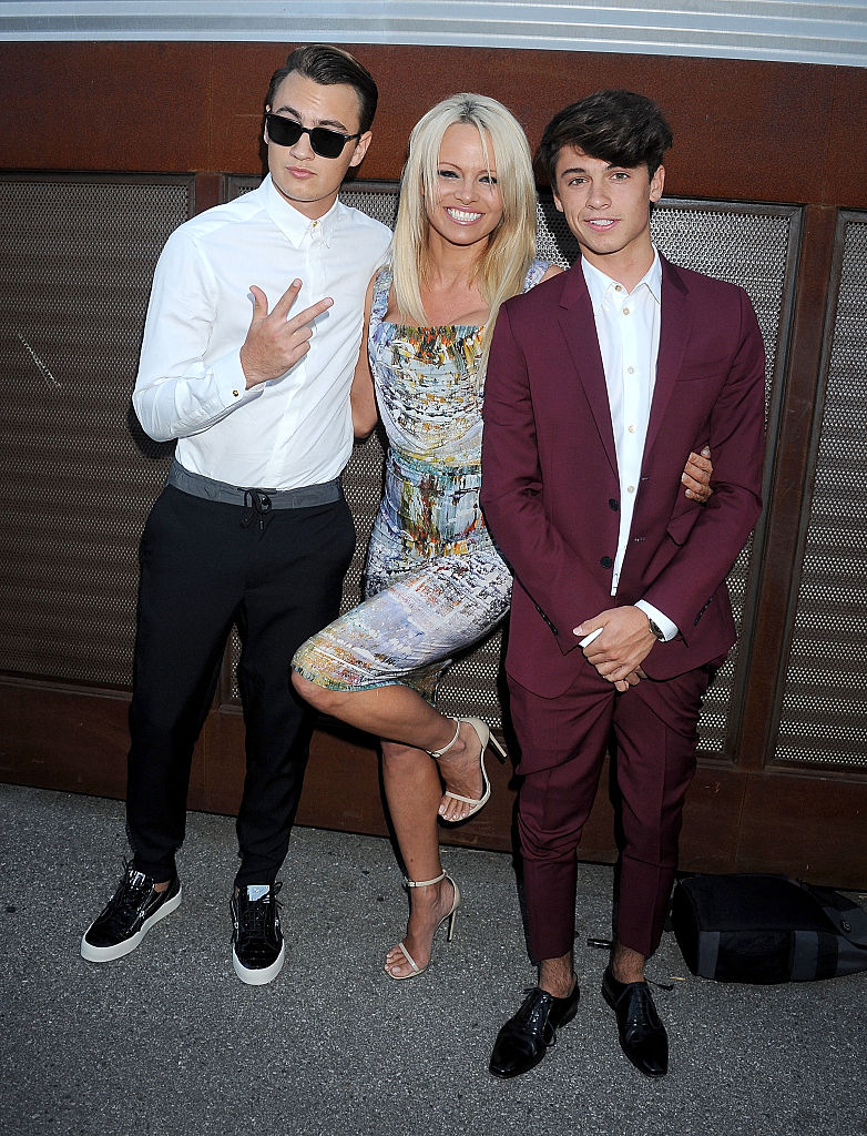Pamelas, Brandon, and Dylan, arrive at The Hidden Heroes Gala presented by Mercy For Animals at Unici Casa on August 29, 2015, in Culver City, California. Brandon wears a white shirt and black pants with sneakers and sunglasses, while Dylan sports a burgundy suit with a white shirt and black loafers. Pamela poses between them in a colorful, fitted dress and strappy heels.