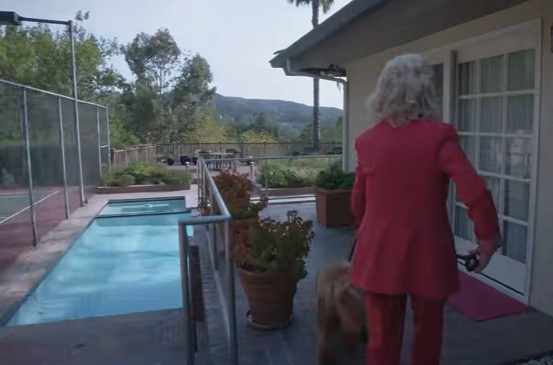 Barbara Eden walks beside the swimming pool toward the glass-paneled doors of her home, accompanied by a small brown dog. Terracotta planters line the paved path, while a fenced tennis court and tree-covered hills can be seen in the distance. | Source: YouTube/People