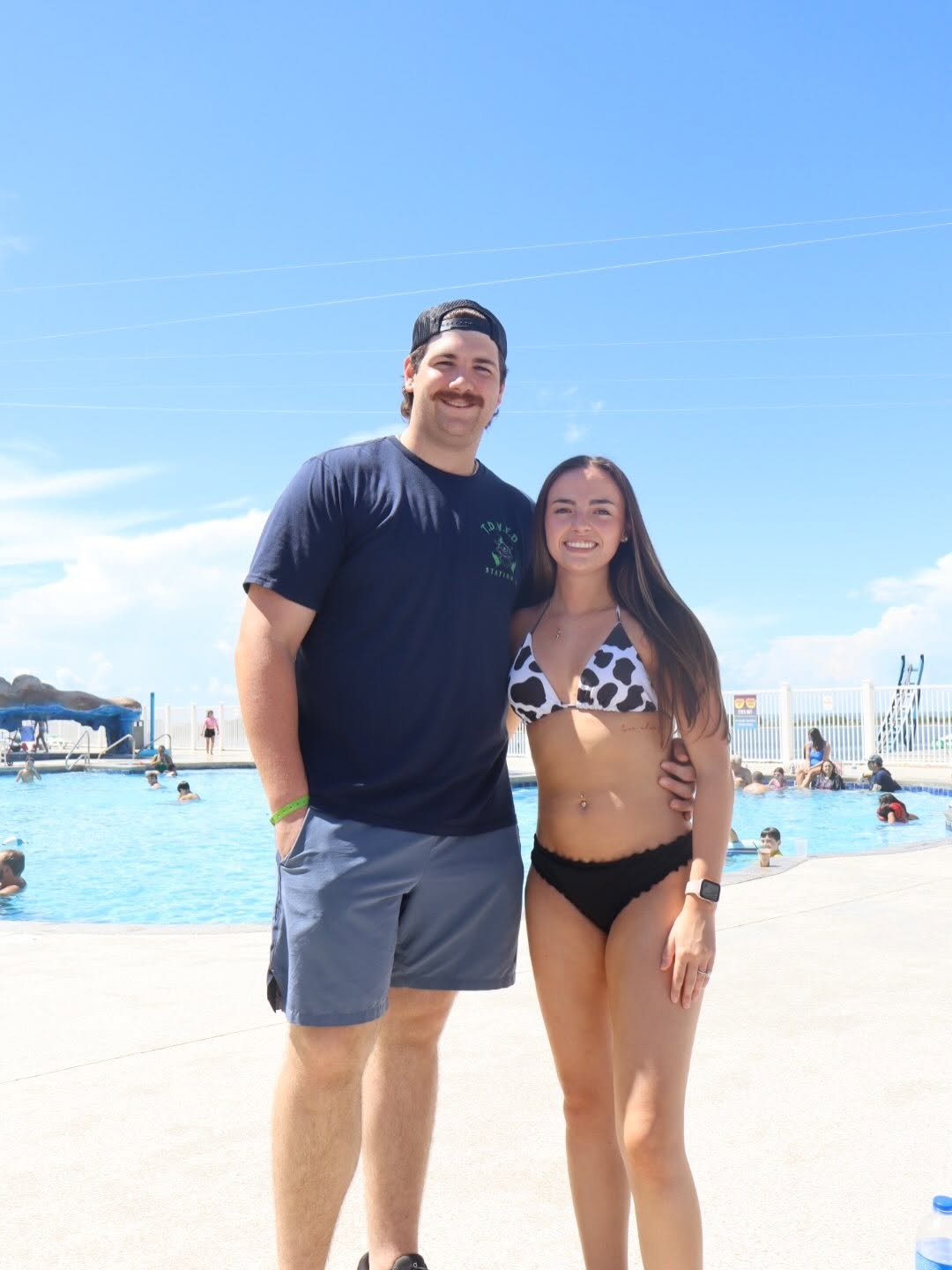 Shane David Hirstius and Leah Victoria Fonte enjoy a sunny day poolside in a sweet summer photo. Shane wraps an arm around Leah while wearing a dark graphic t-shirt, casual grey shorts, and a backwards black cap, as Leah smiles alongside him in a black-and-white cow-print bikini top with black bottoms under a bright blue sky. | Source: Facebook/Shane Hirstius