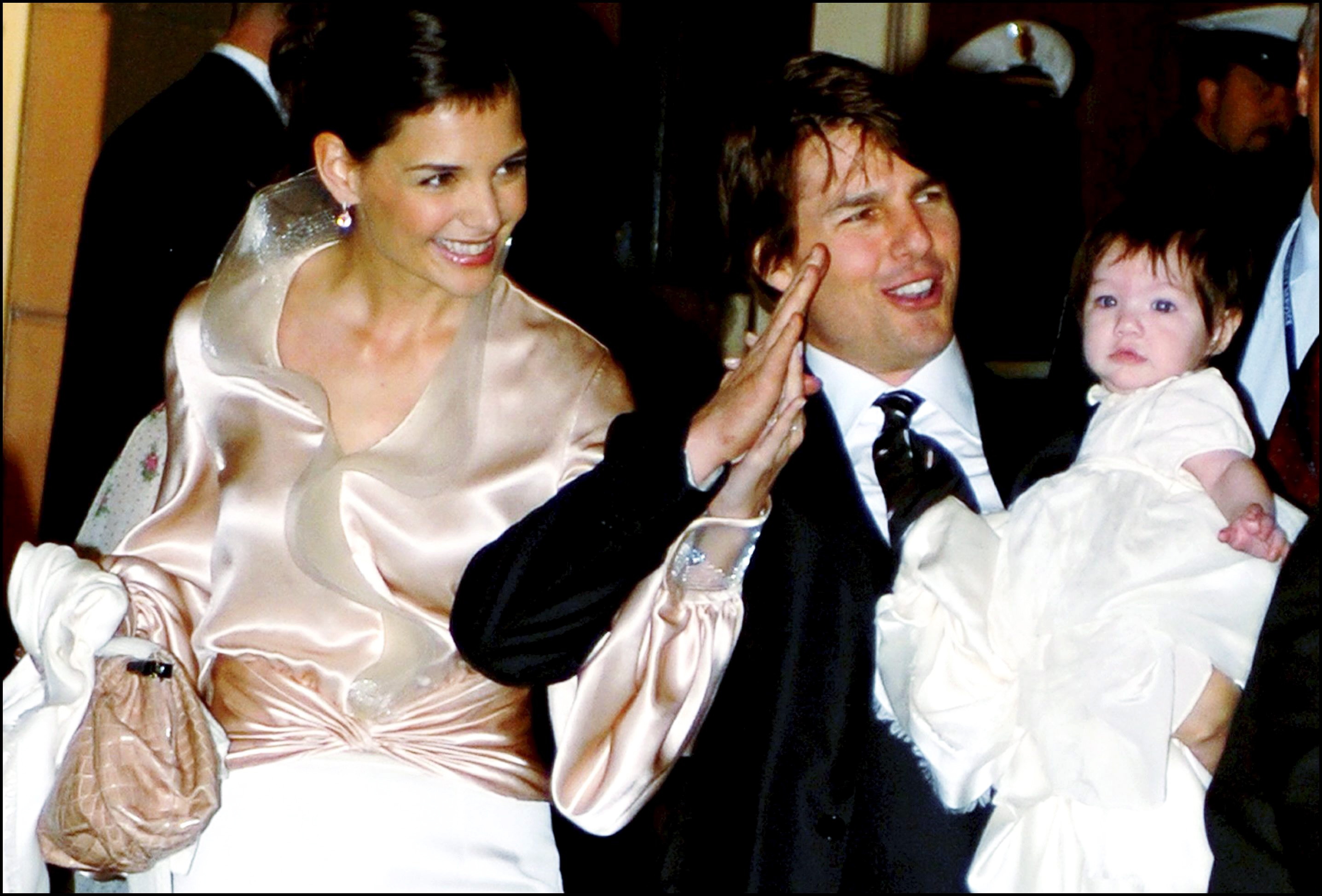 In this nostalgic snapshot, a baby Suri Cruise is cradled in her father's arms, nestled securely as her parents — Katie Holmes and Tom Cruise — make a rare public appearance together. Smiling and holding hands, the couple moves through the crowd with ease, their faces lit up as they acknowledge onlookers. It’s a moment that captures both the height of their stardom and the early days of their journey as a family.
