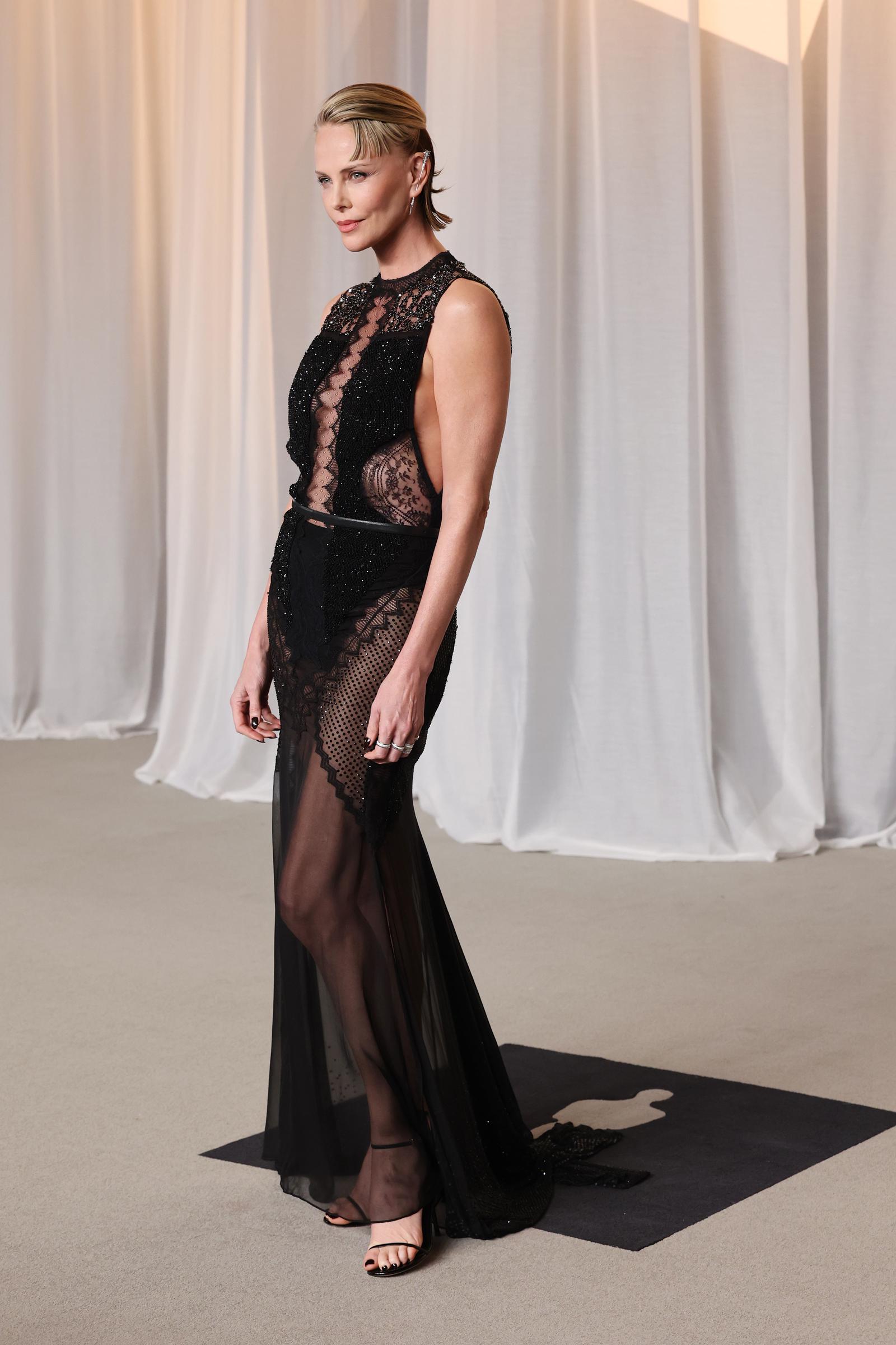 Charlize Theron brings fearless drama to the red carpet in a sleeveless sheer black gown intricately layered with delicate lace, beadwork, and fishnet panels. Accented by a slim waist belt and styled with a sleek, wet-look updo and minimalist black sandals, the Oscar winner pairs edgy modern glamour with classic movie-star poise as she celebrates receiving the Icon Award.