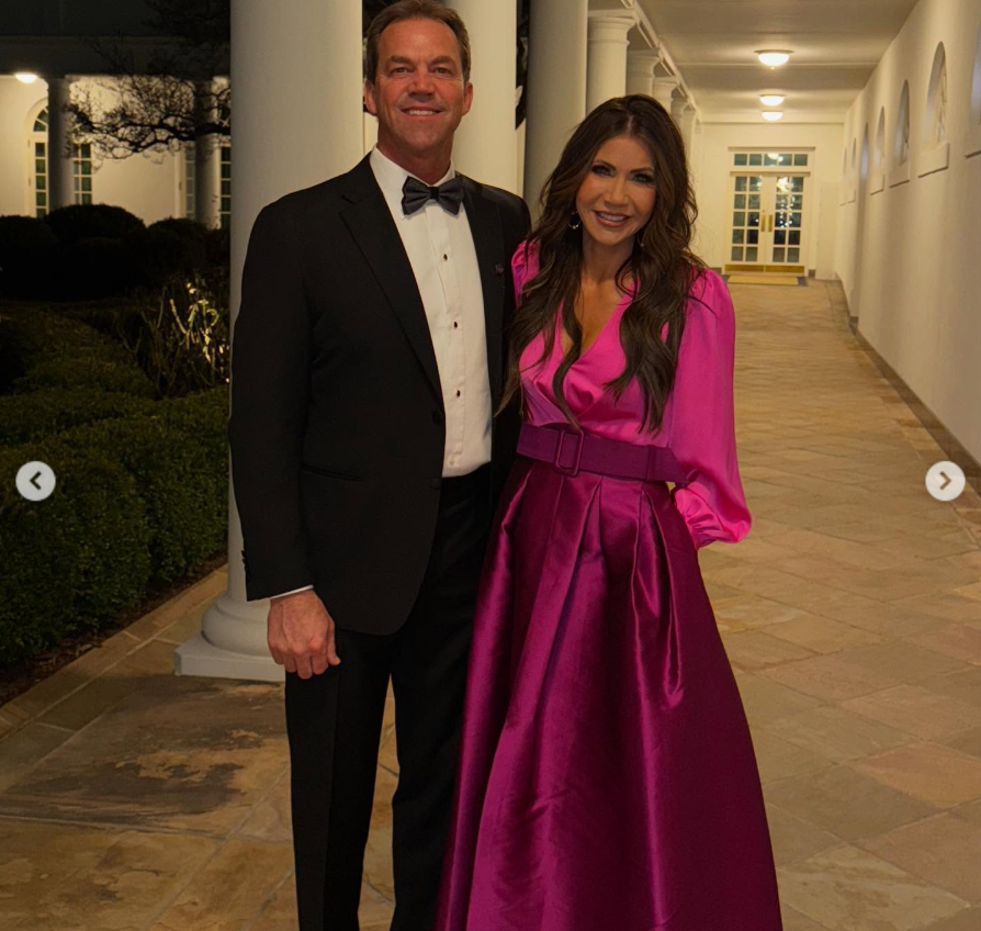 Kristi and Bryon Noem pose together at night along a column-lined walkway, smiling for the camera in a softly lit outdoor setting. | Source: Instagram/kristinoem