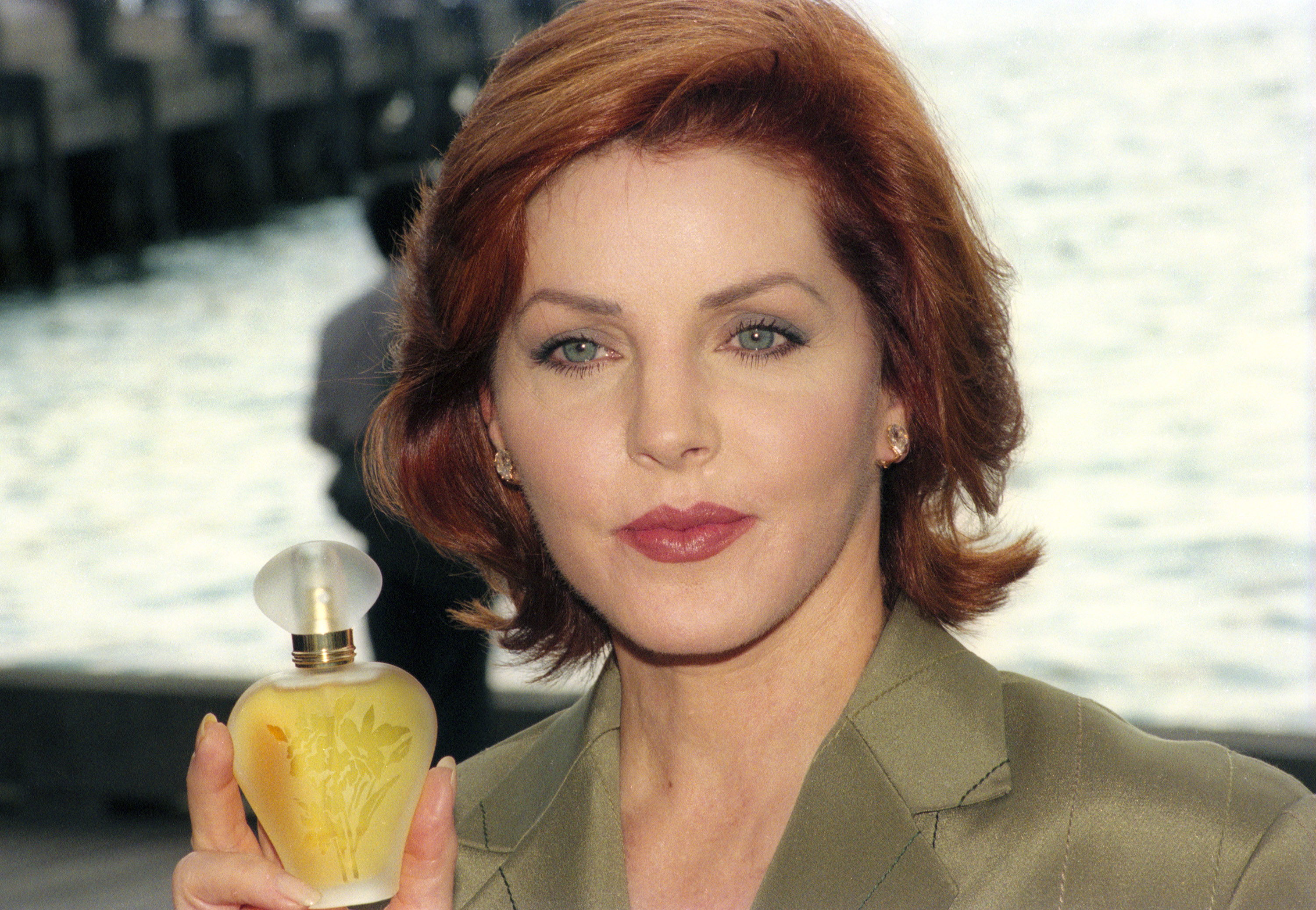 Priscilla Presley holds up a frosted glass bottle of her fragrance "Moments" during its launch at the Park Hyatt Hotel in Sydney, Australia, on October 29, 1996, wearing an olive-green tailored jacket and styling her vibrant auburn hair in a soft, flipped-out bob. | Source: Getty Images