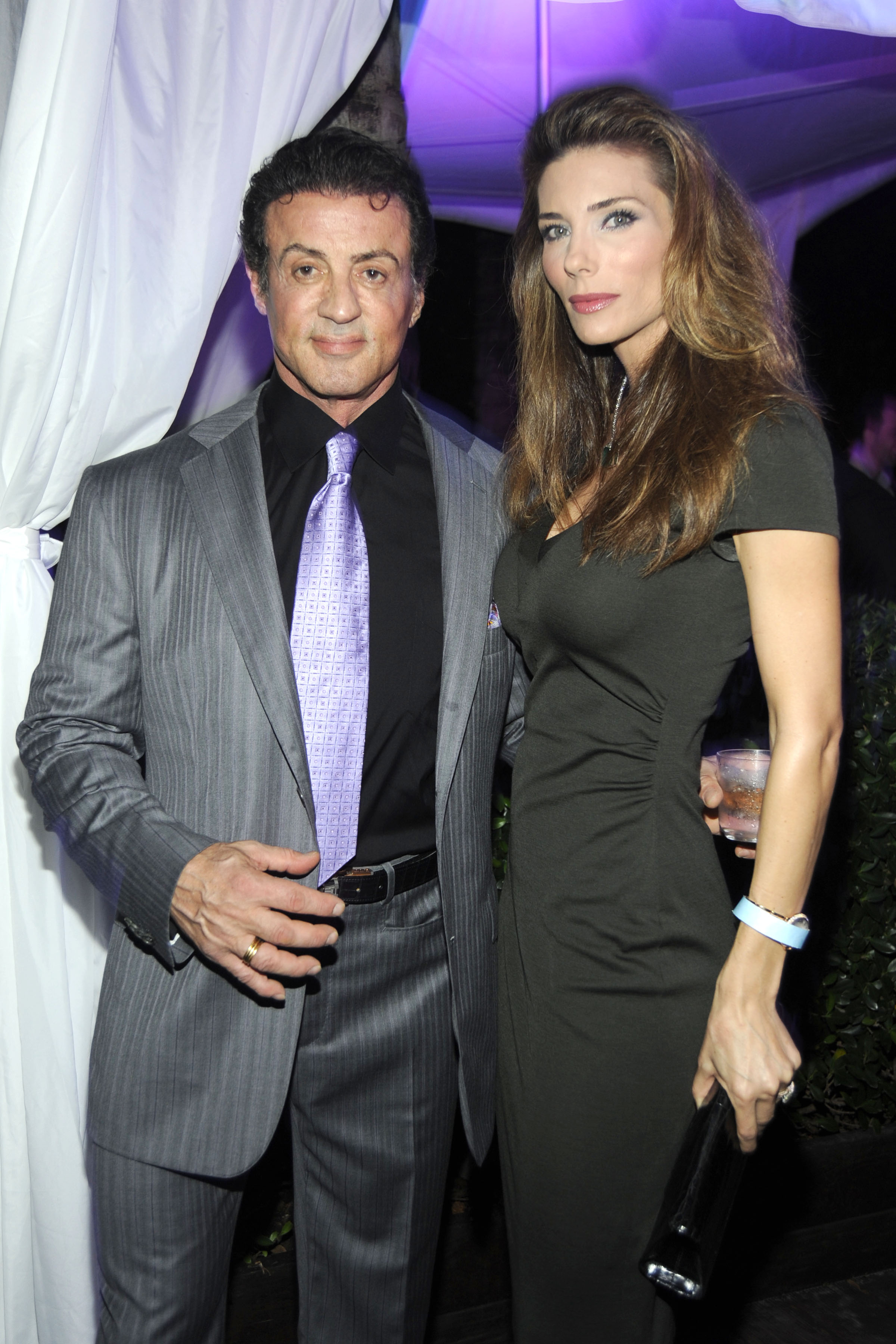 Sylvester Stallone and Jennifer Flavin attended a Galerie Gmurzynska dinner at The Setai in Miami Beach, Florida, on December 2, 2009. Posing beneath purple lighting, Stallone wore a gray pinstripe suit with a lavender tie, while Flavin chose a fitted black dress and carried a clutch. The appearance captured the longtime spouses 12 years into their marriage, as they continued building the family life that ultimately spanned decades together.