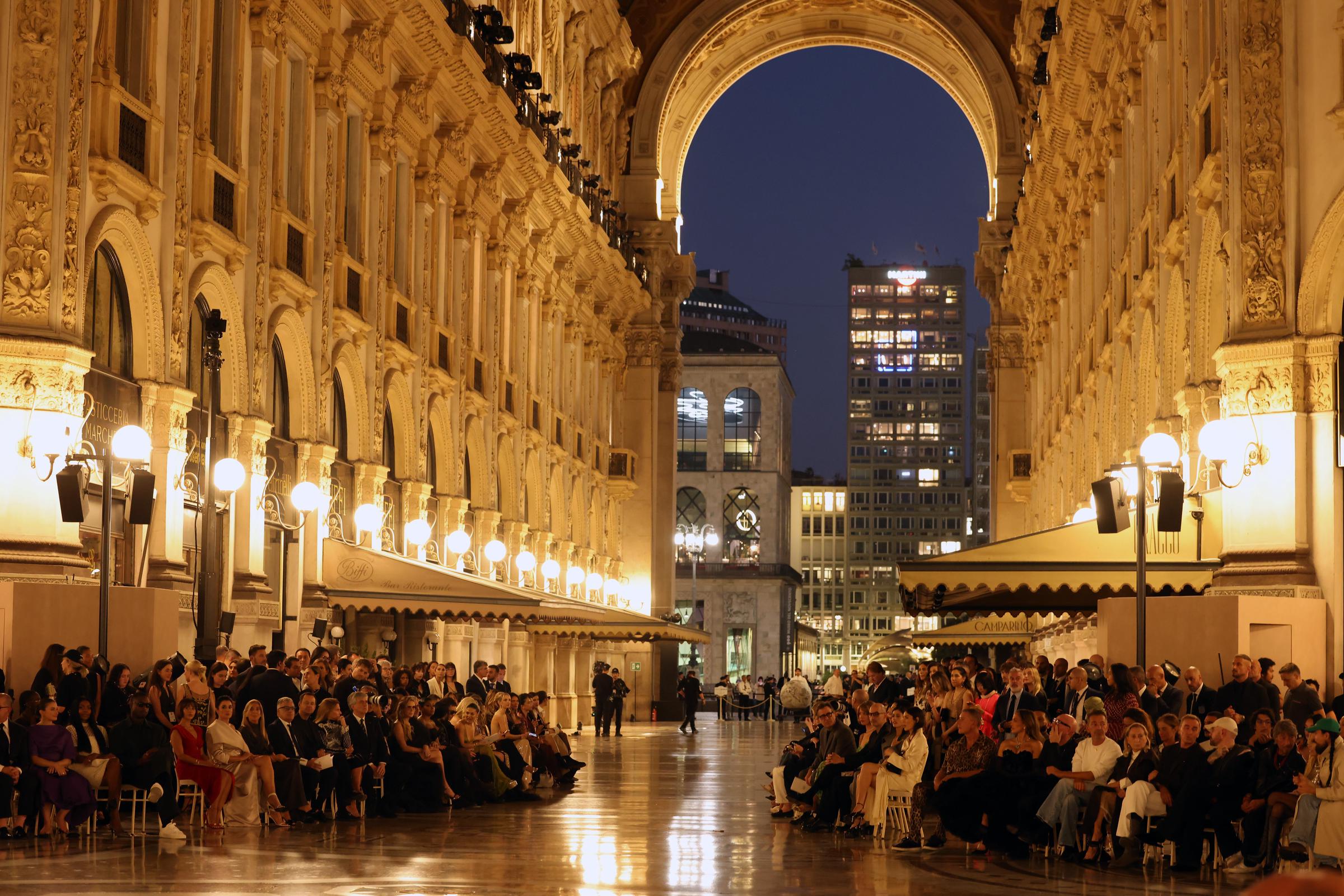 Galleria Vittorio Emanuele II transformed into a breathtaking open-air catwalk for Vogue World: Milano 2026 on September 22, 2026, in Milan, Italy. Glowing street lamps illuminated rows of seated fashion spectators along the polished stone corridor, framing a dramatic view through the towering historic archway toward the illuminated city skyline beyond. | Source: Getty Images