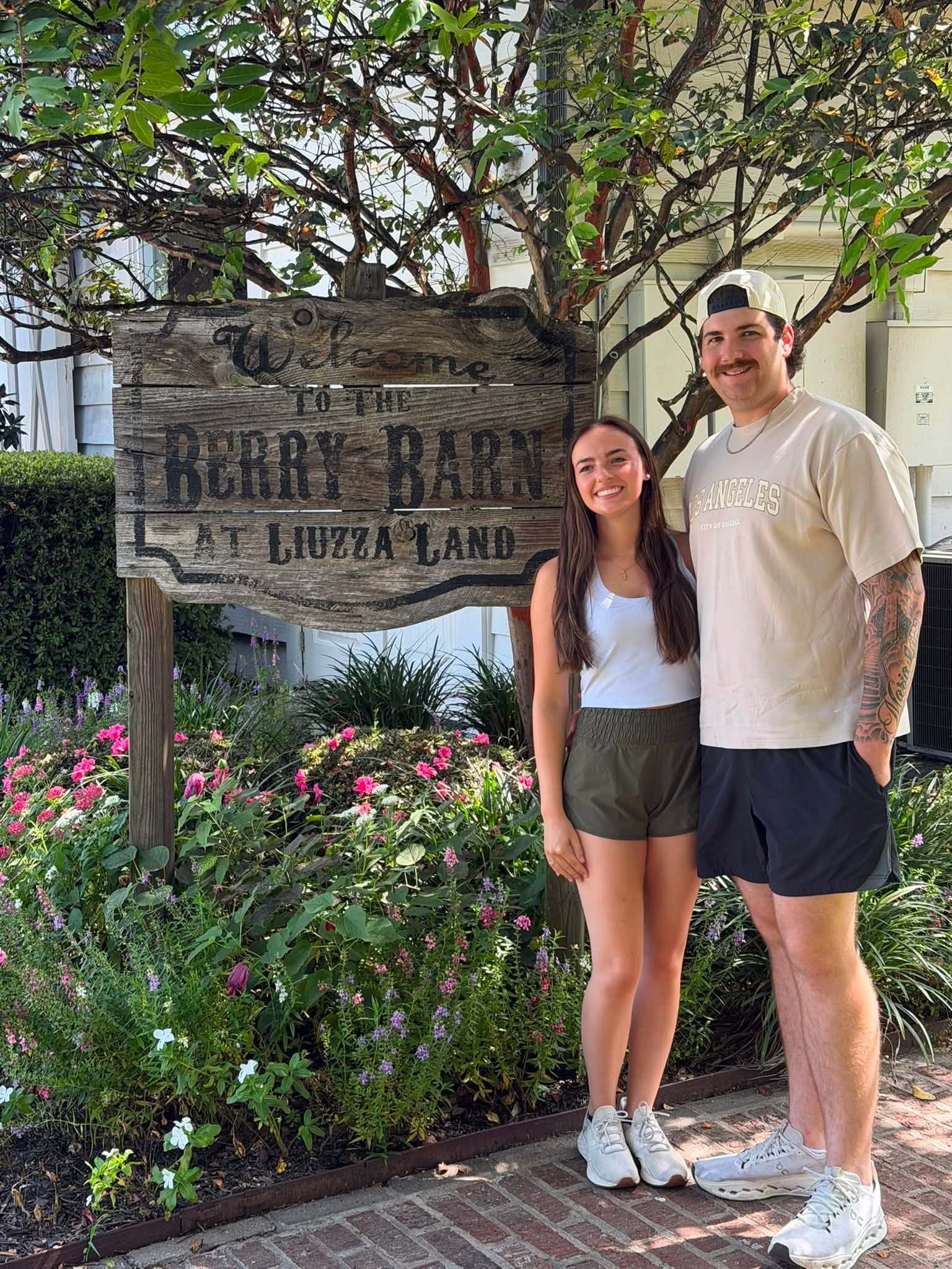 Leah Victoria Fonte and Shane David Hirstius pose together during an outing at The Berry Barn at Liuzza Land in an upbeat snapshot shared by Shane's parents. Leah smiles in a white tank top, olive green athletic shorts, and white sneakers, standing beside Shane, who smiles in a beige graphic t-shirt, dark shorts, white athletic shoes, and a backwards cap in front of lush blooming flowers and a rustic wooden welcome sign. | Source: Facebook/StevenMarjorie Hirstius