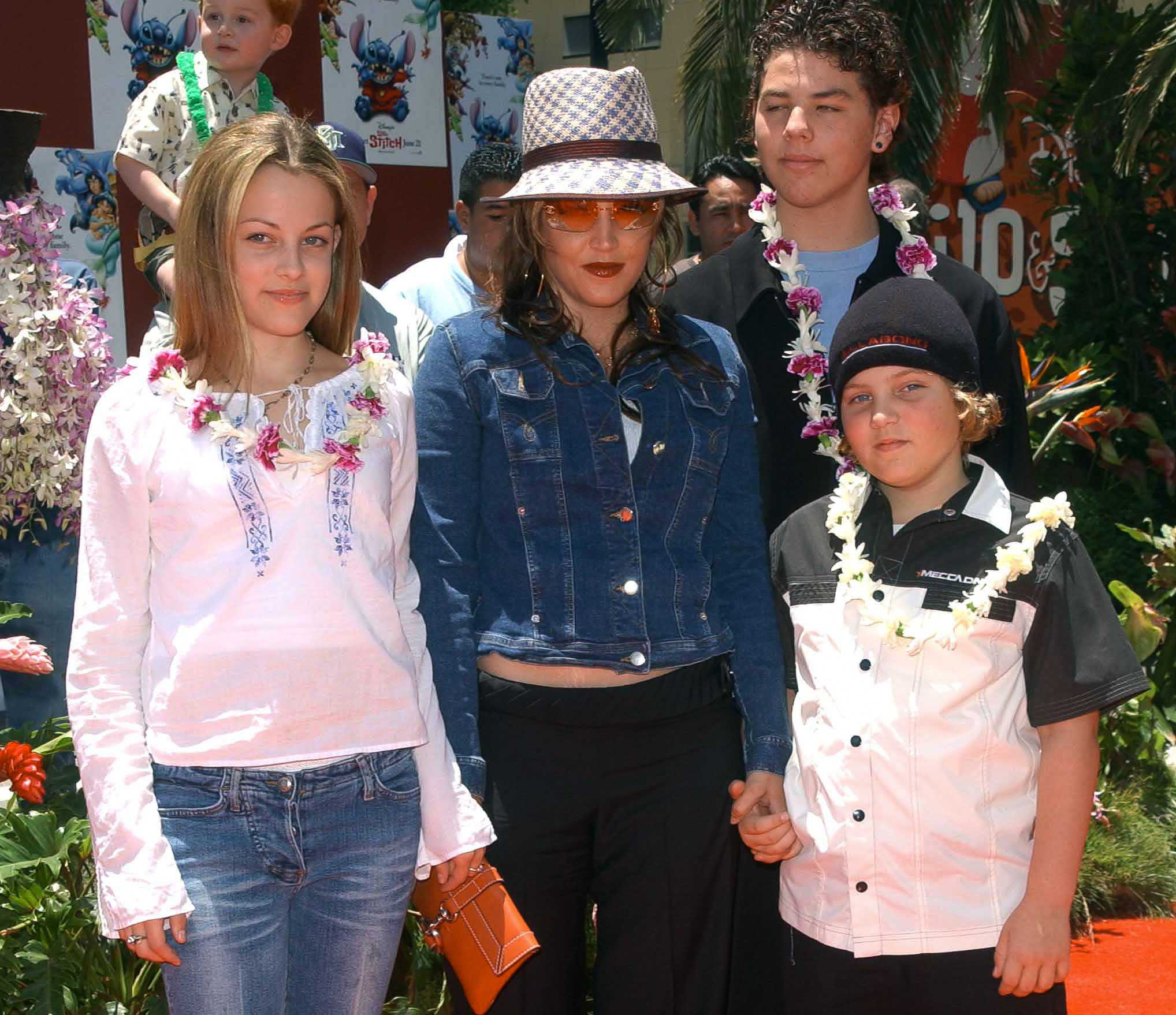 Lisa Marie Presley poses with her children Riley Keough and Benjamin Keough, alongside her half-brother Navarone Garibaldi, at the premiere of "Lilo and Stitch" at the El Capitan Theatre in Hollywood, California, on June 16, 2002, with Presley wearing a denim jacket, tinted sunglasses, and a patterned fedora, while the family embraces Hawaiian leis on the red carpet.