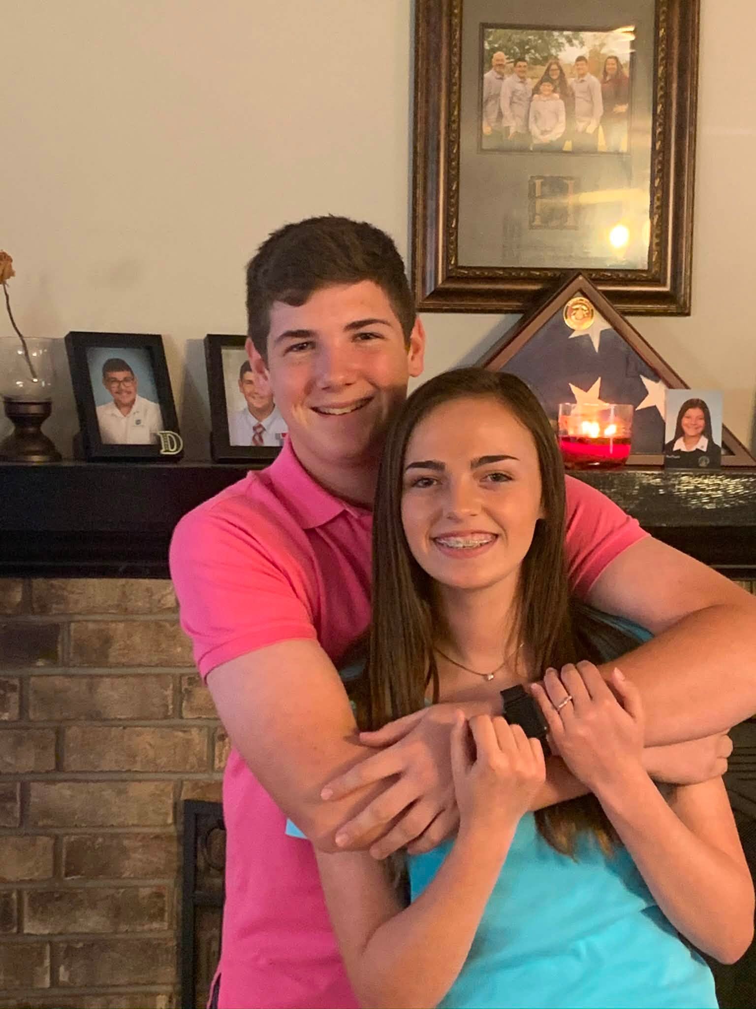 Shane David Hirstius and Leah Victoria Fonte share a youthful hug in an early snapshot from their high school days together. Shane wraps his arms around Leah from behind wearing a bright pink polo shirt, while Leah smiles with braces in a turquoise top against a brick fireplace mantel lined with framed family photos. | Source: Facebook/StevenMarjorie Hirstius