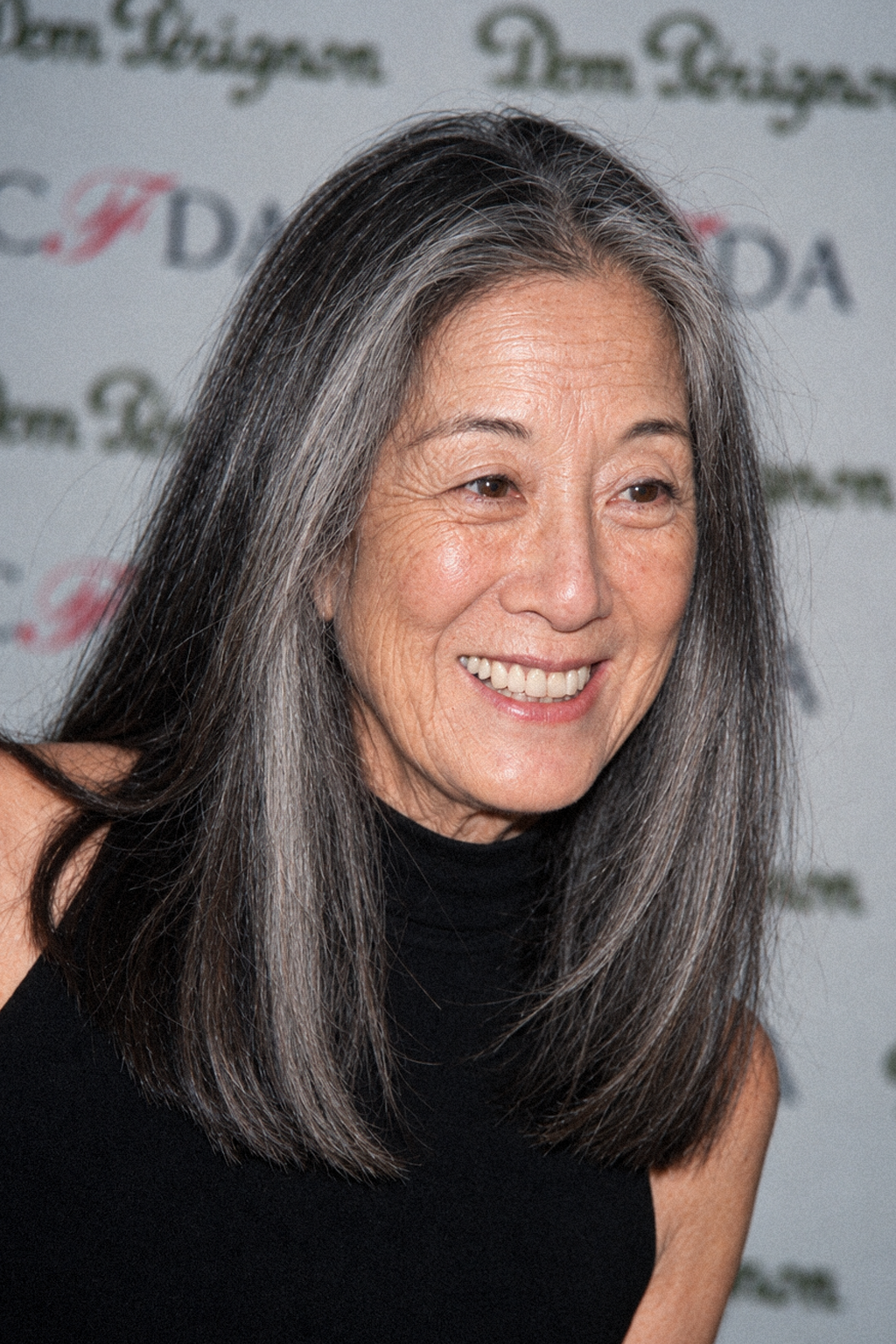 Pictured against an event step-and-repeat backdrop, this AI rendering presents Vera Wang flashing a broad smile, showing off authentic mature skin texture, salt-and-pepper shoulder-length hair, and her natural pre-rhinoplasty bridge. Styled in a sleek black halter-neck silhouette, the speculative image captures the celebrated designer embracing an untouched aging journey. | Source: ChatGPT