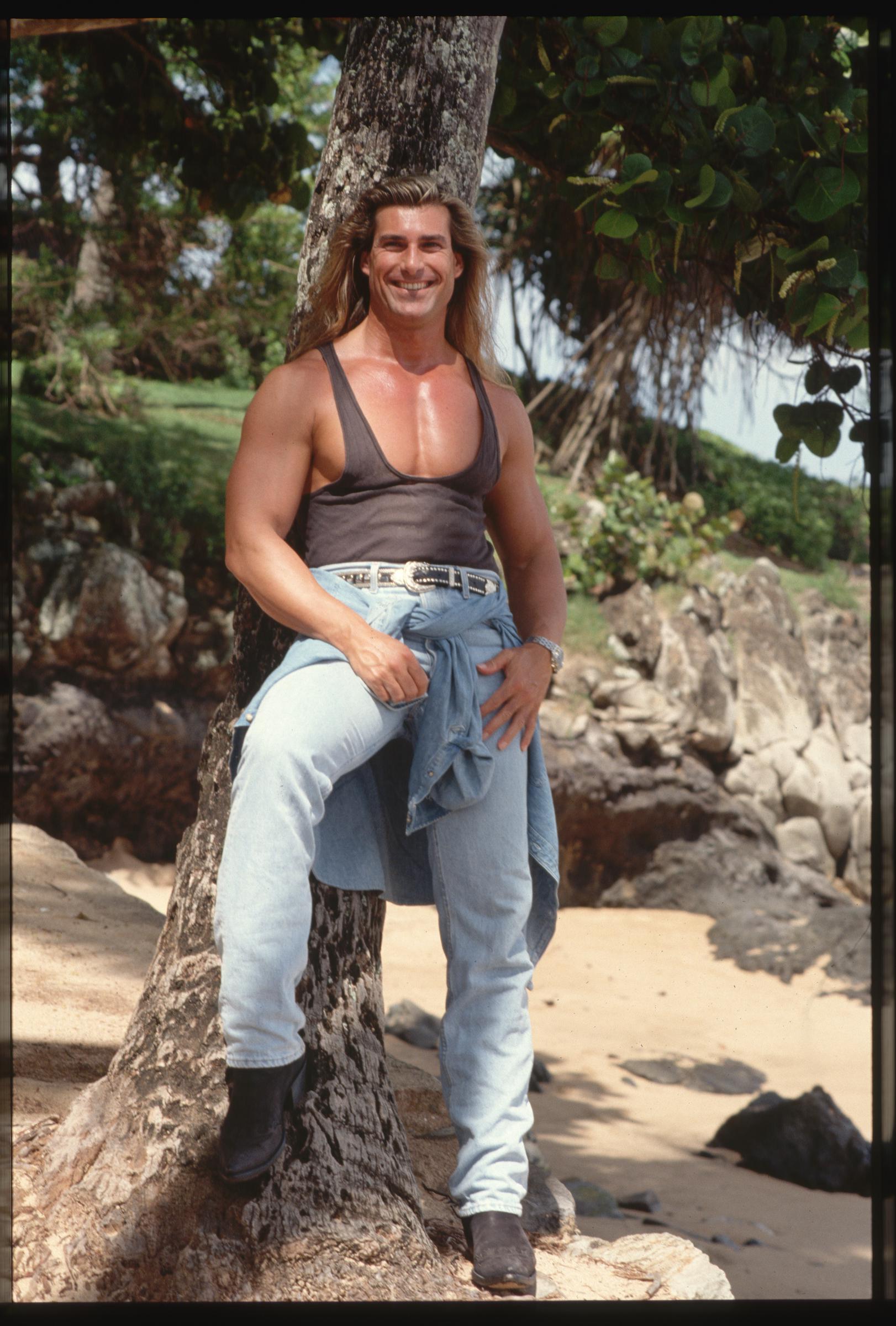 At first, he had no idea he was on any book covers. In this photo, the iconic romance book cover star wore a tank top and jeans while leaning against a tree in Hawaii on January 1, 1993. | Source: Getty Images